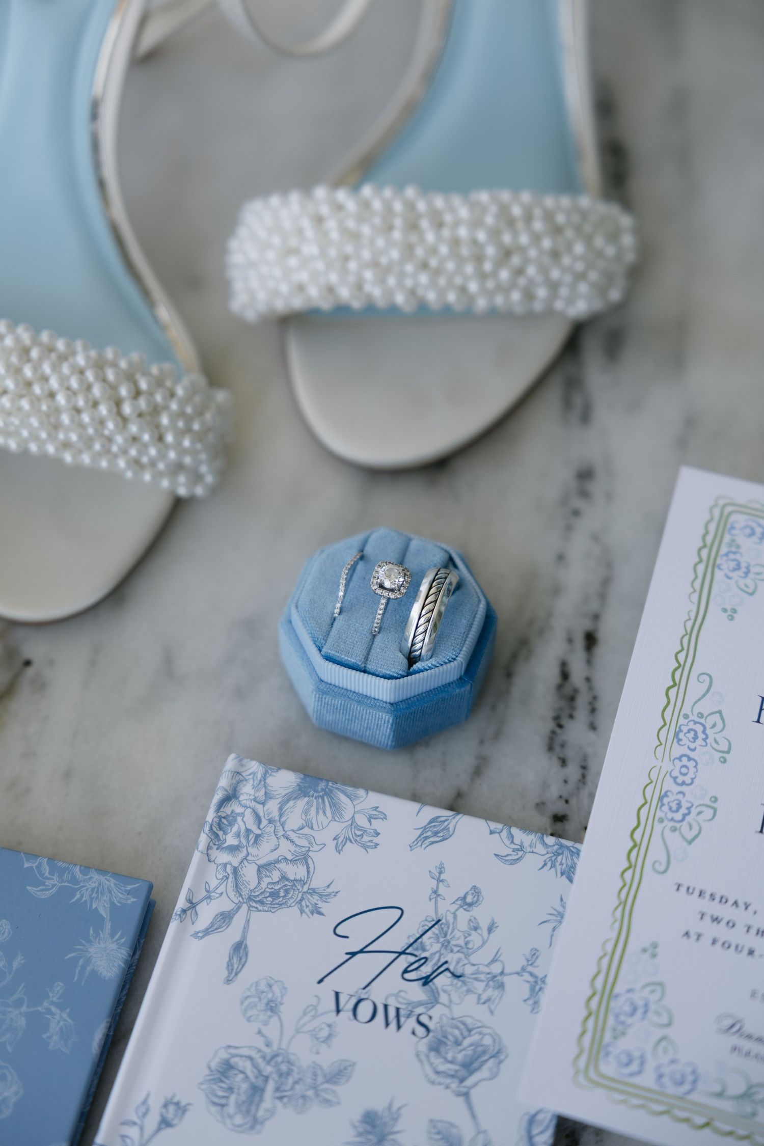 Blue velvet ring box with diamond wedding rings beside pearl bridal heels, vow book, and wedding invitation on marble.