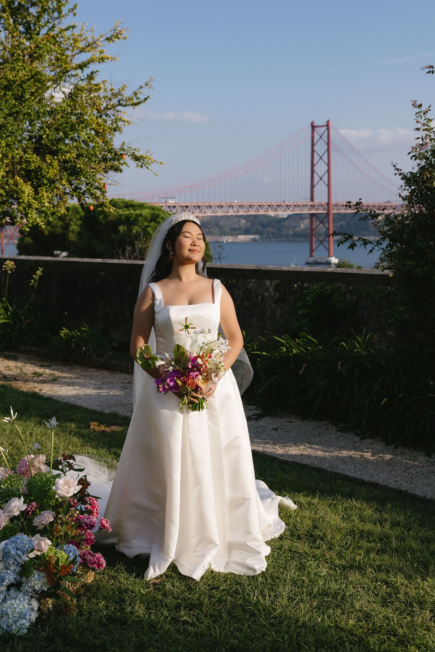 Bride in white gown holding colorful bouquet stands in garden with iconic red suspension bridge and river in background.