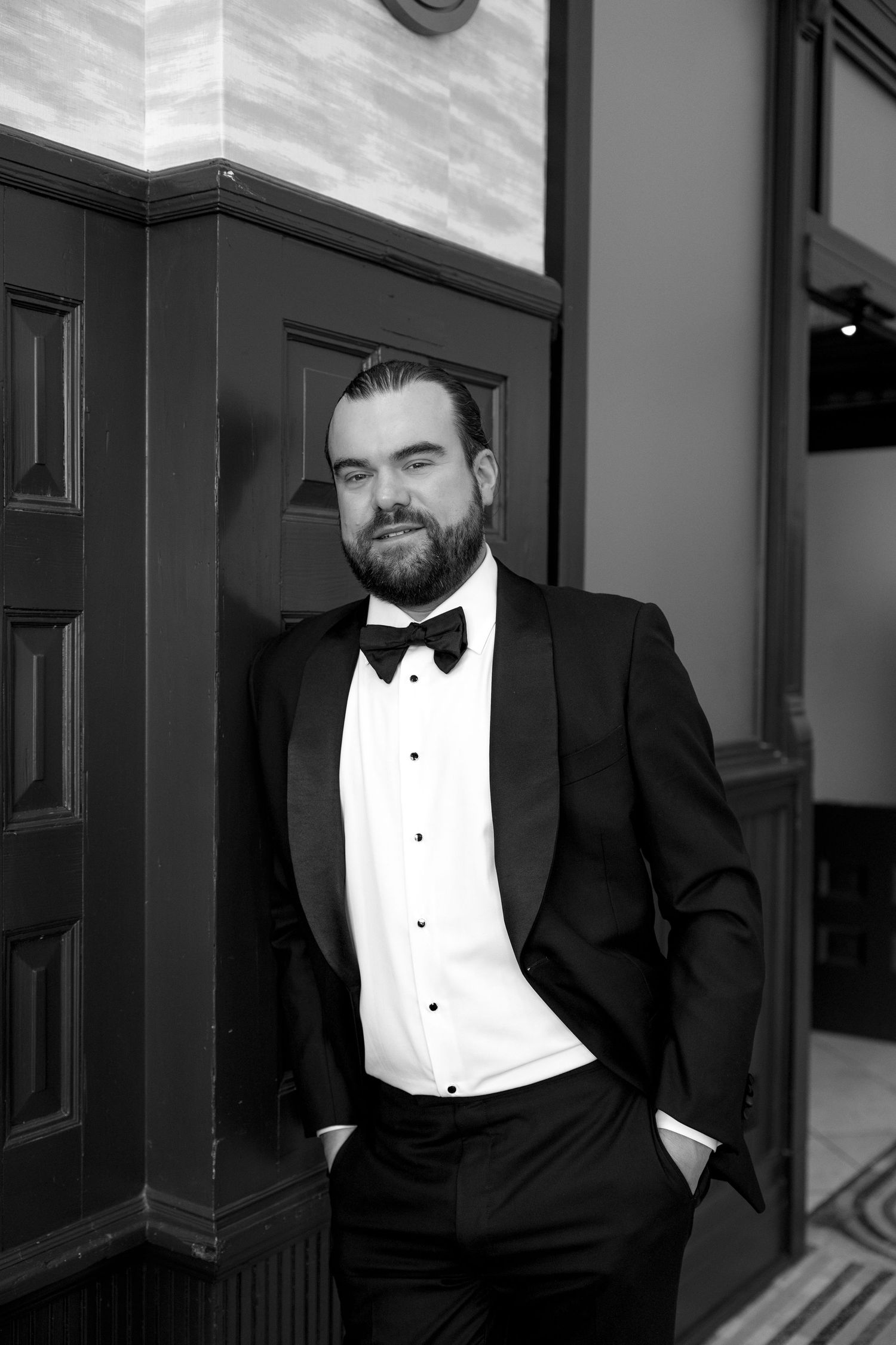 Bearded man in black tuxedo with bow tie leaning against wooden door in black and white portrait photo.