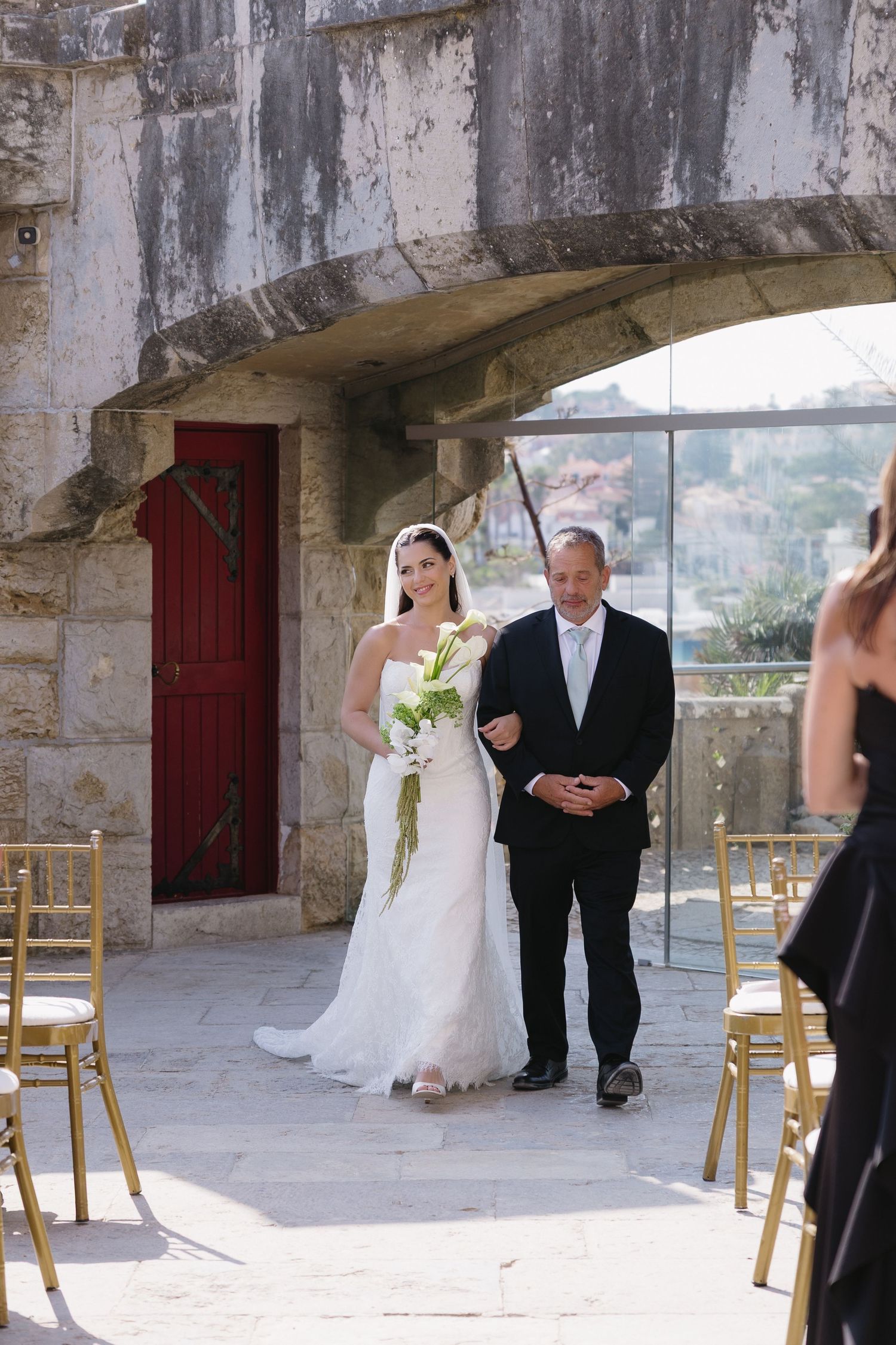 Bride in white gown walks down outdoor aisle with escort at stone venue wedding ceremony.