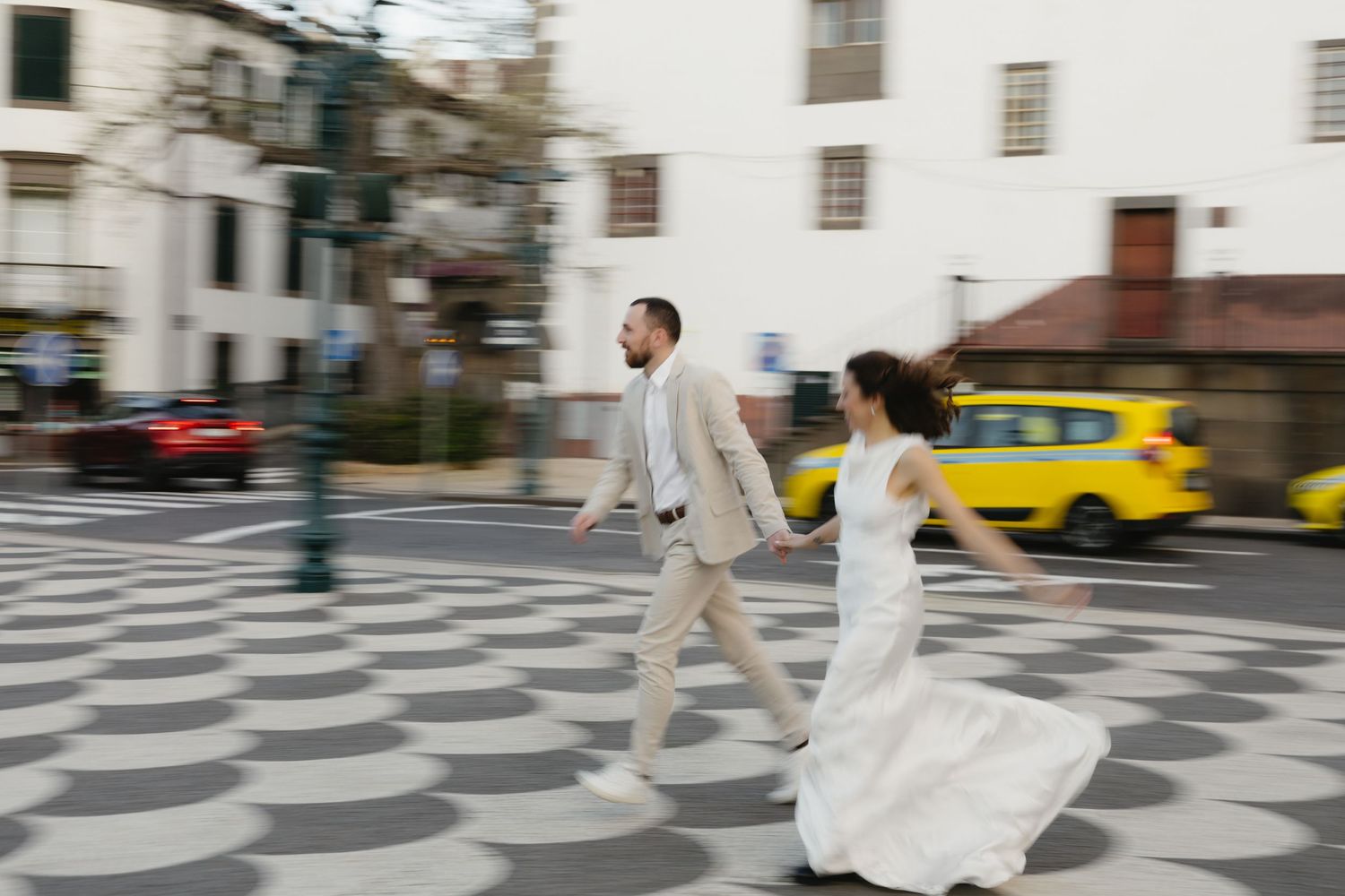 Bride and groom cross a cobblestone street in motion blur with a yellow taxi passing nearby.