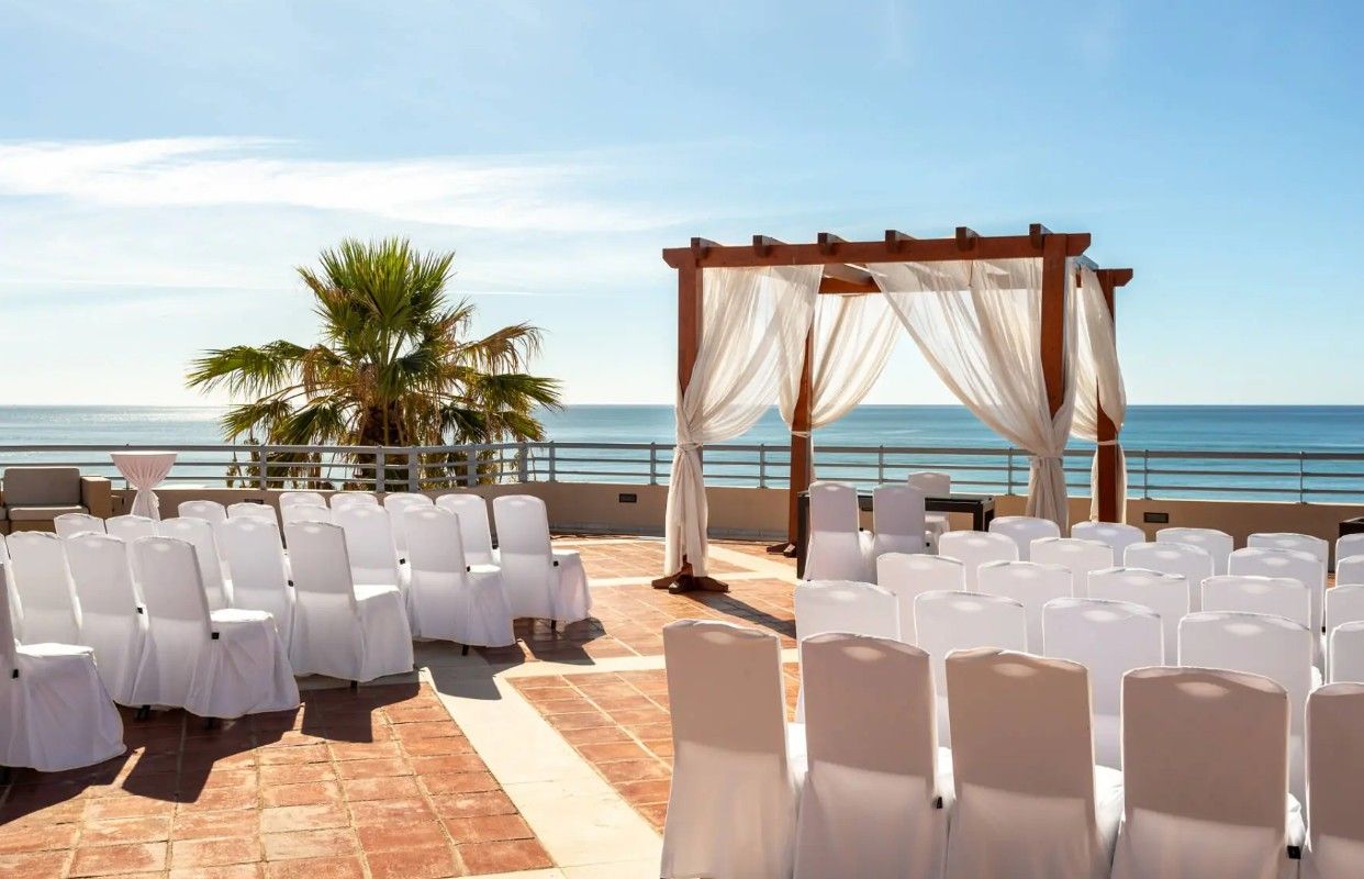Outdoor beachfront wedding ceremony with white draped pergola, white chairs, and palm tree backdrop.