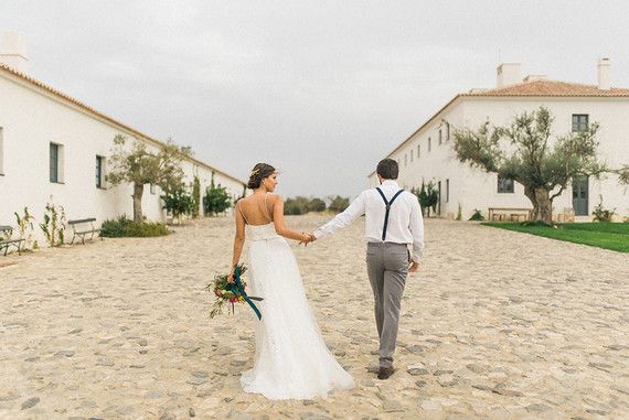 Bride and groom walking hand-in-hand across a cobblestone courtyard at a white Portuguese wedding estate.