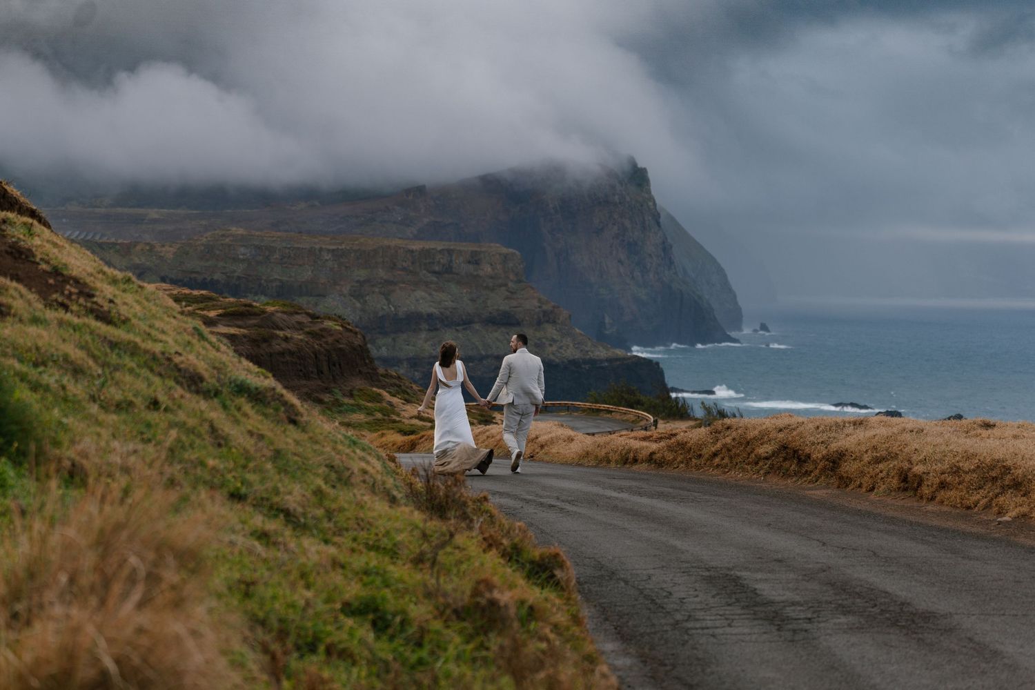 Bride and groom walking hand in hand along a dramatic coastal road.