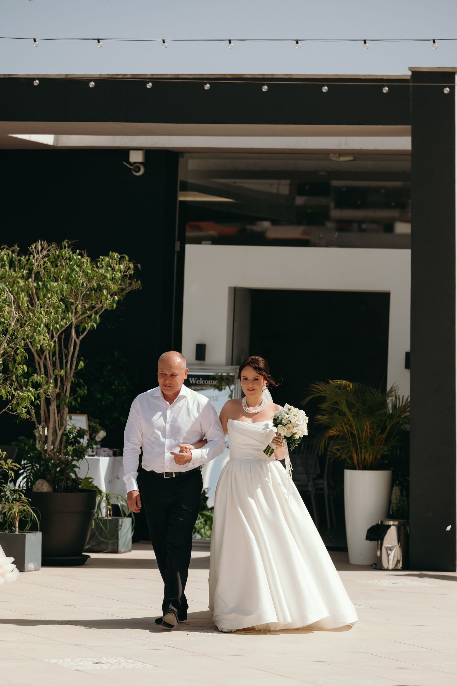 Bride in white strapless gown walks with escort holding white bouquet at outdoor wedding venue.