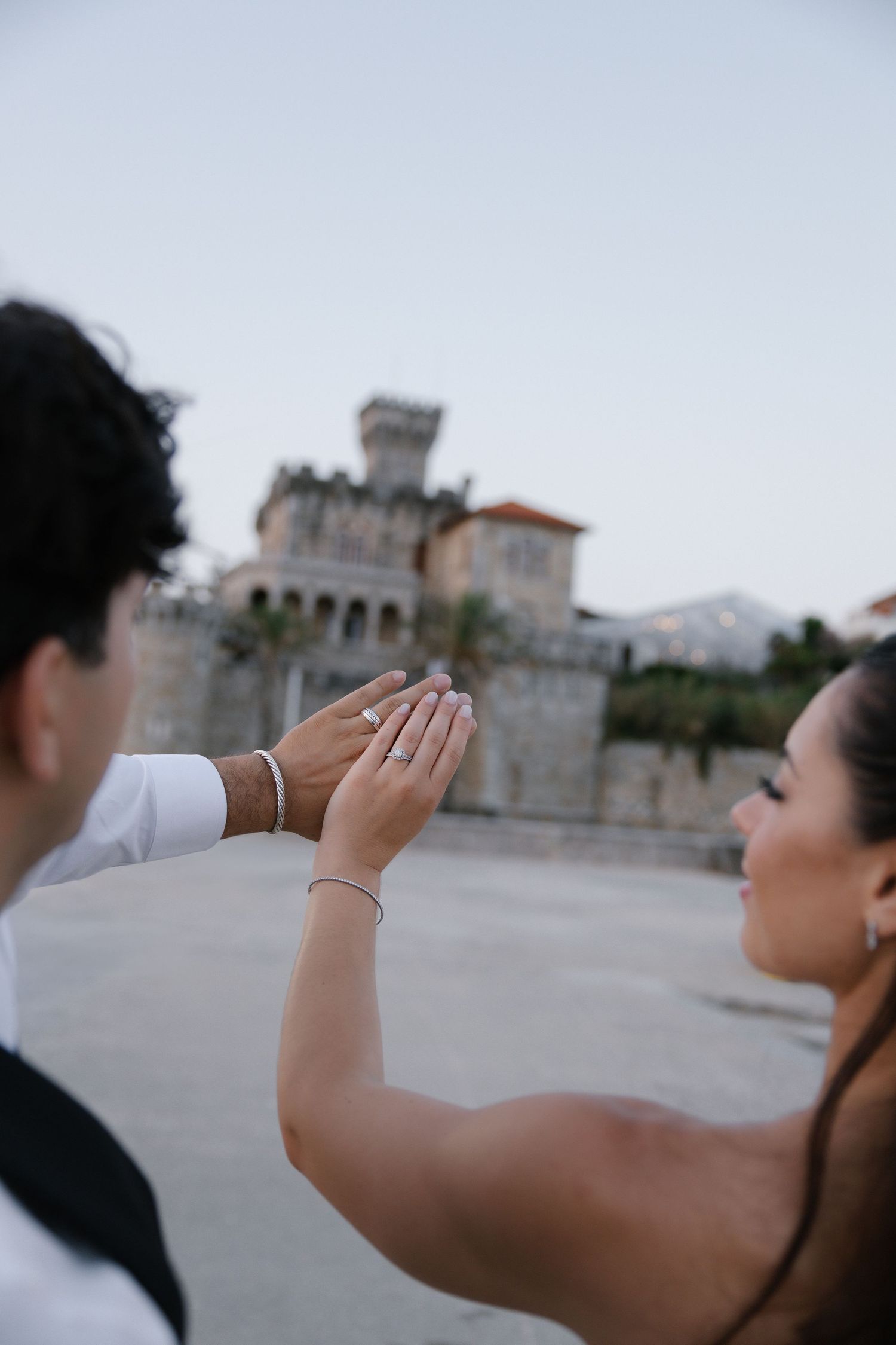 Couple holding hands showing engagement ring with a castle in the background during an outdoor photo shoot.