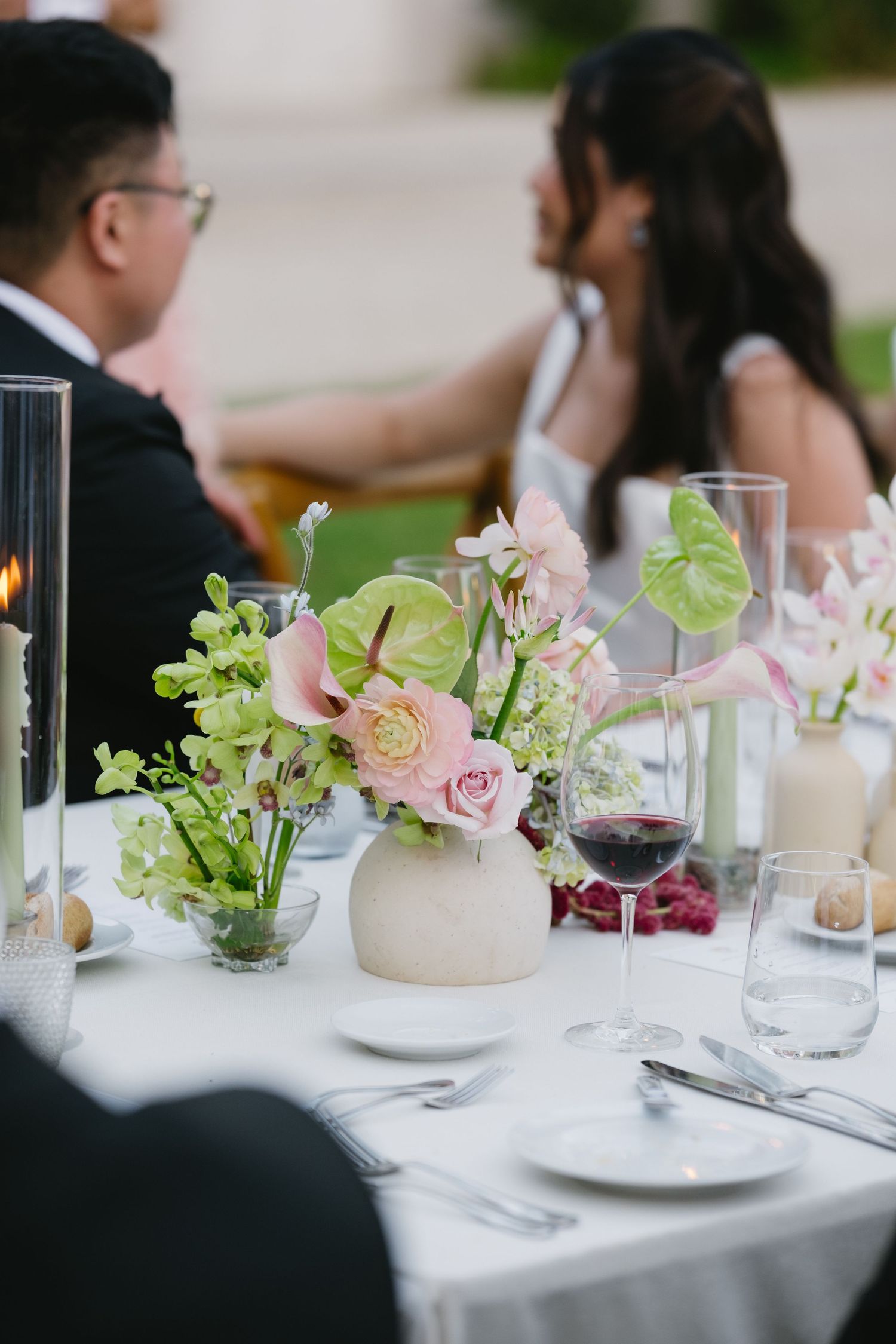 Elegant wedding reception table with pink floral centerpiece, wine glass, and candles, guests seated in background.