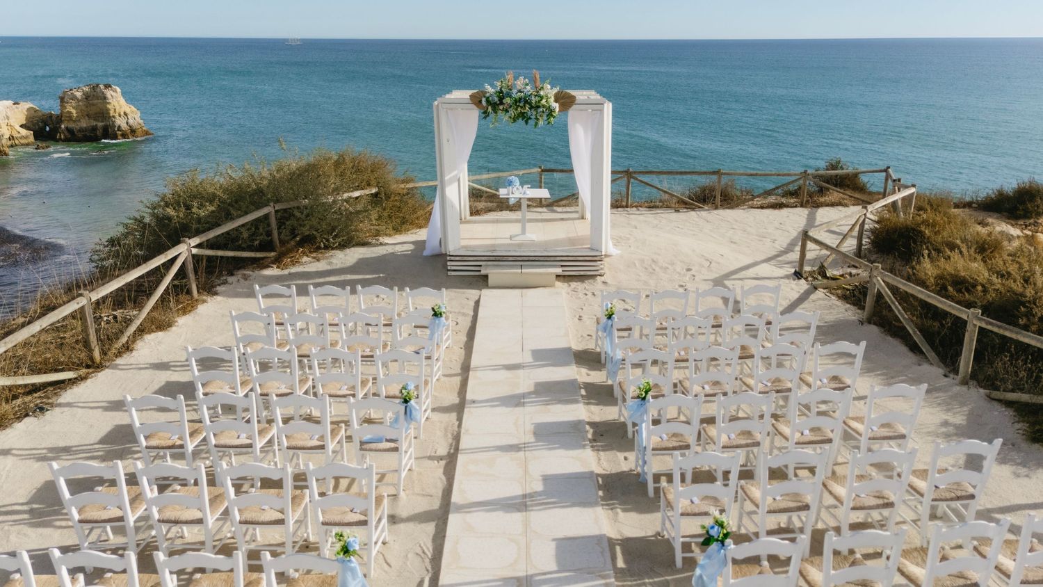 Elegant beachfront wedding ceremony setup with white chairs and floral arch overlooking the turquoise sea.