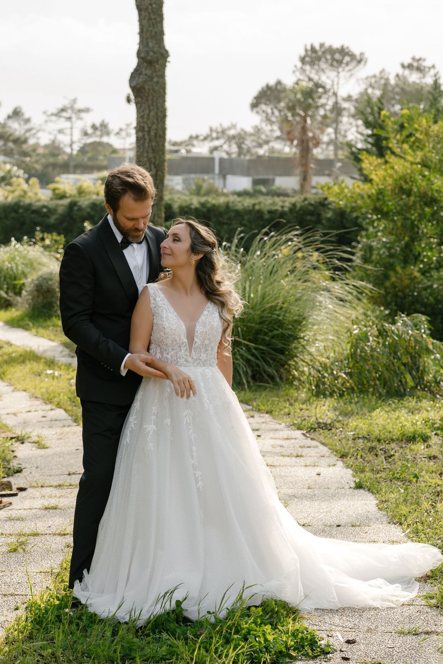 Bride in lace ball gown and groom in black tuxedo posing on garden pathway.