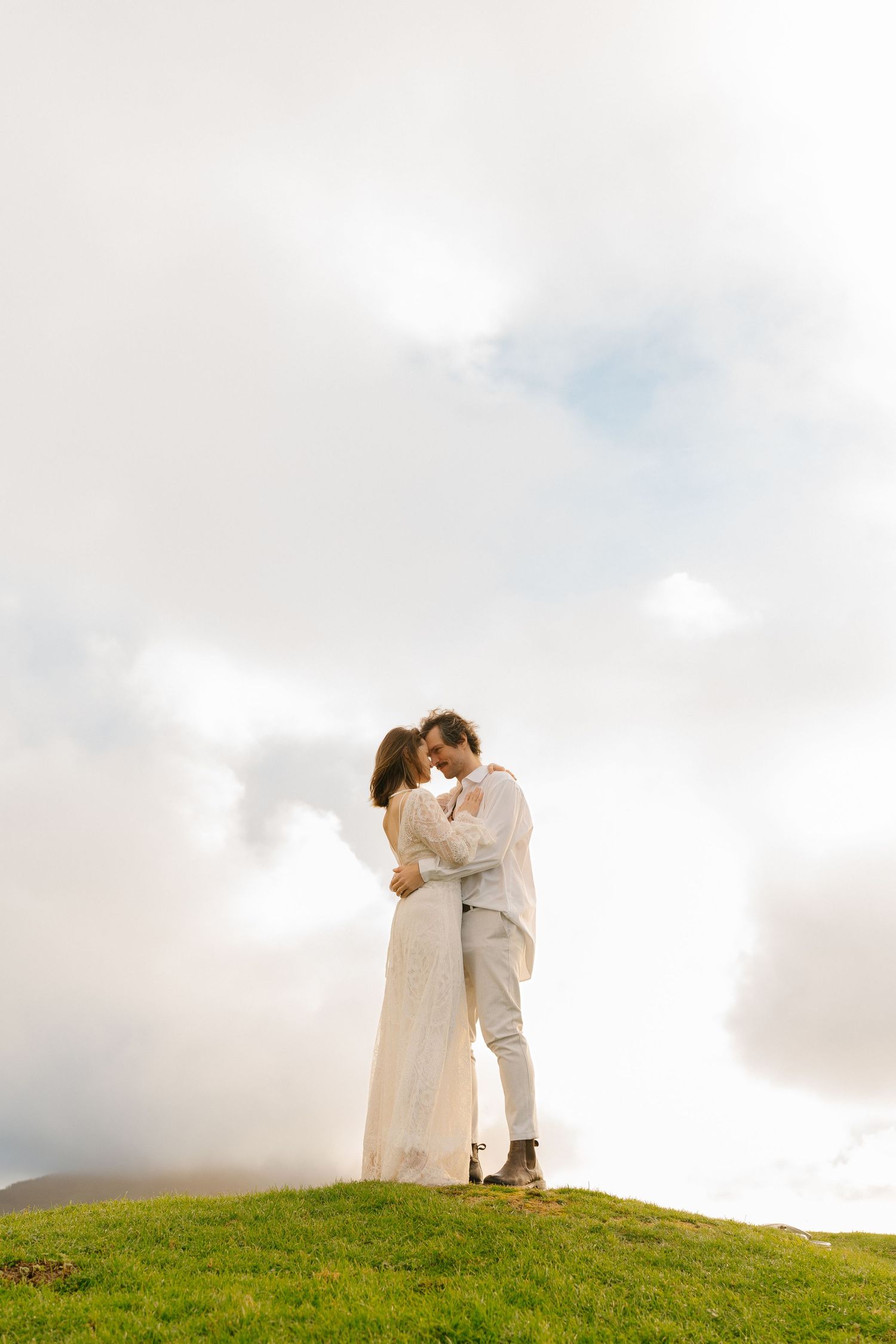 Couple embracing on a misty green hilltop, bride in white lace gown and groom in light suit against cloudy sky.
