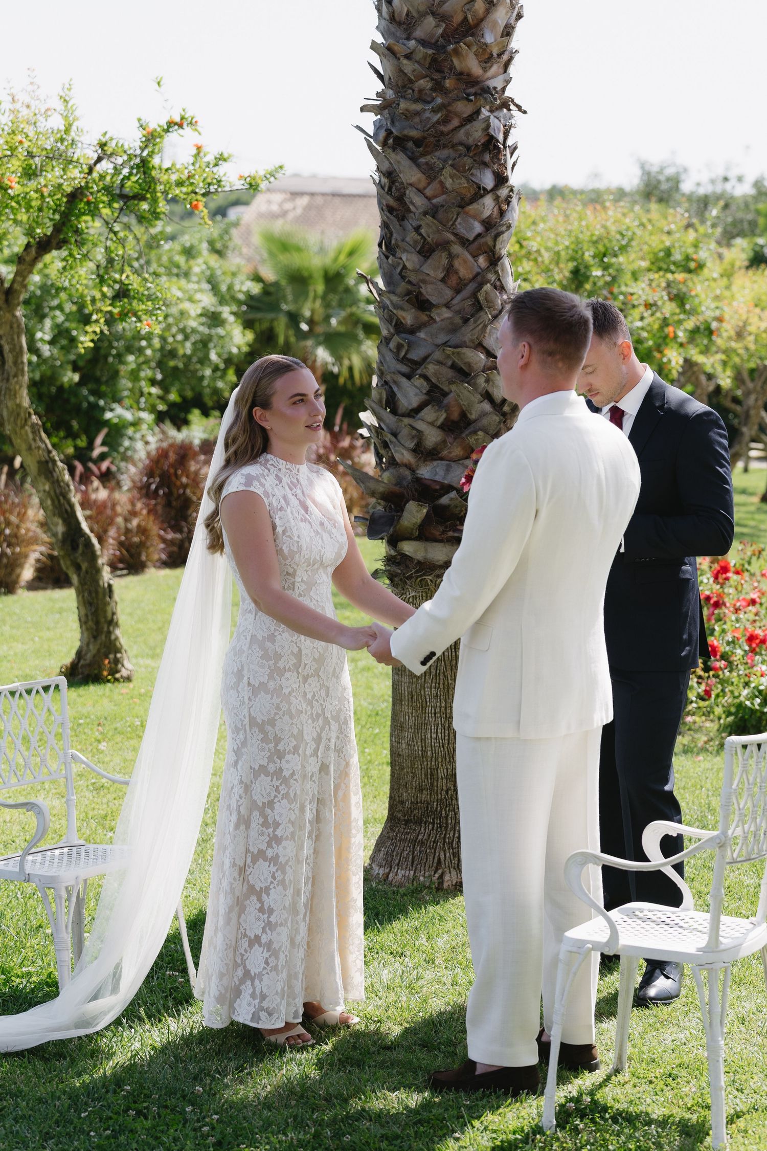 Bride and groom exchange vows outdoors under palm tree, officiant present, white garden chairs nearby.