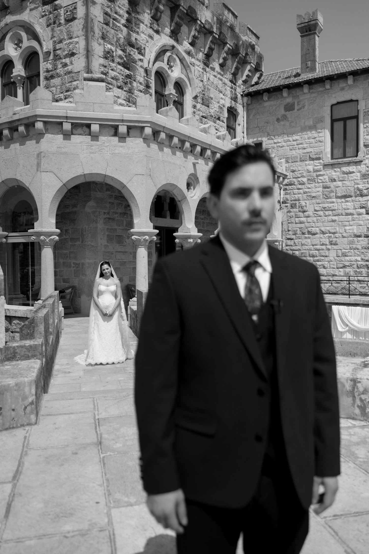 Black and white wedding photo of groom in suit with bride in white dress posed before historic stone building.