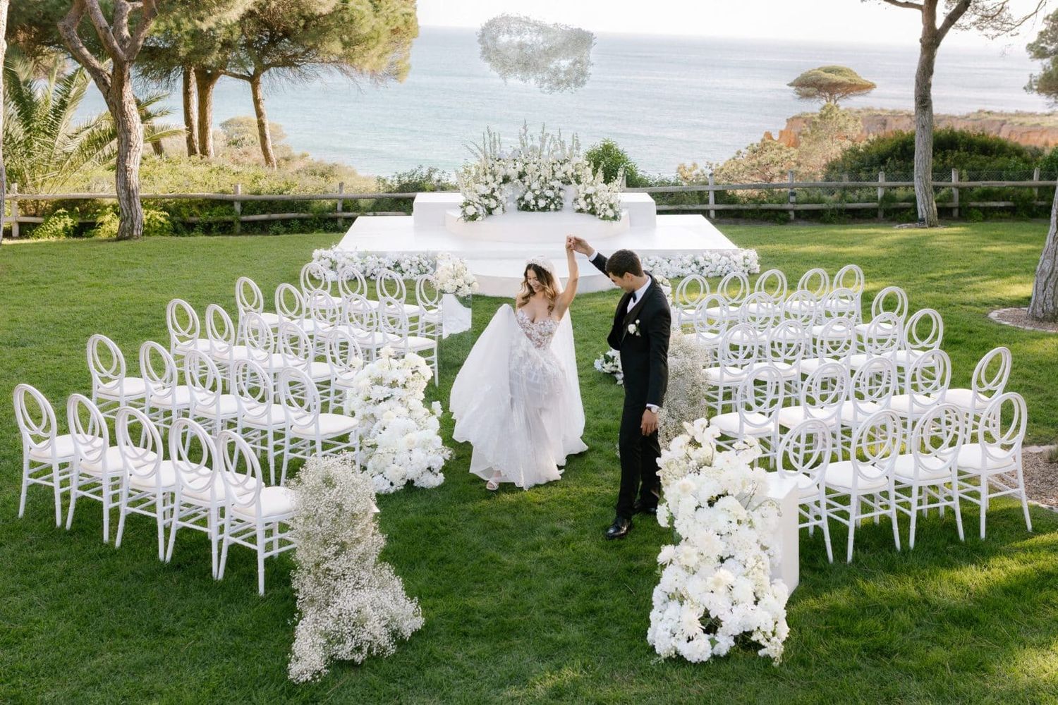 Couple dancing at elegant all-white outdoor wedding ceremony setup with floral arrangements and white chairs on green lawn.