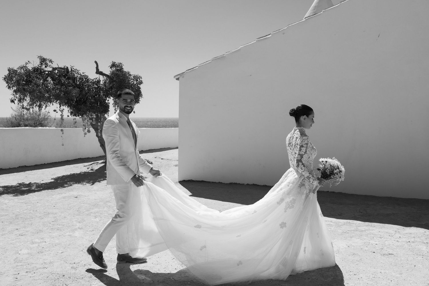 Wedding couple walking together at Nossa Senhora chapel in Algarve, bride in a white dress and groom in a suit, surrounded by cliffs.