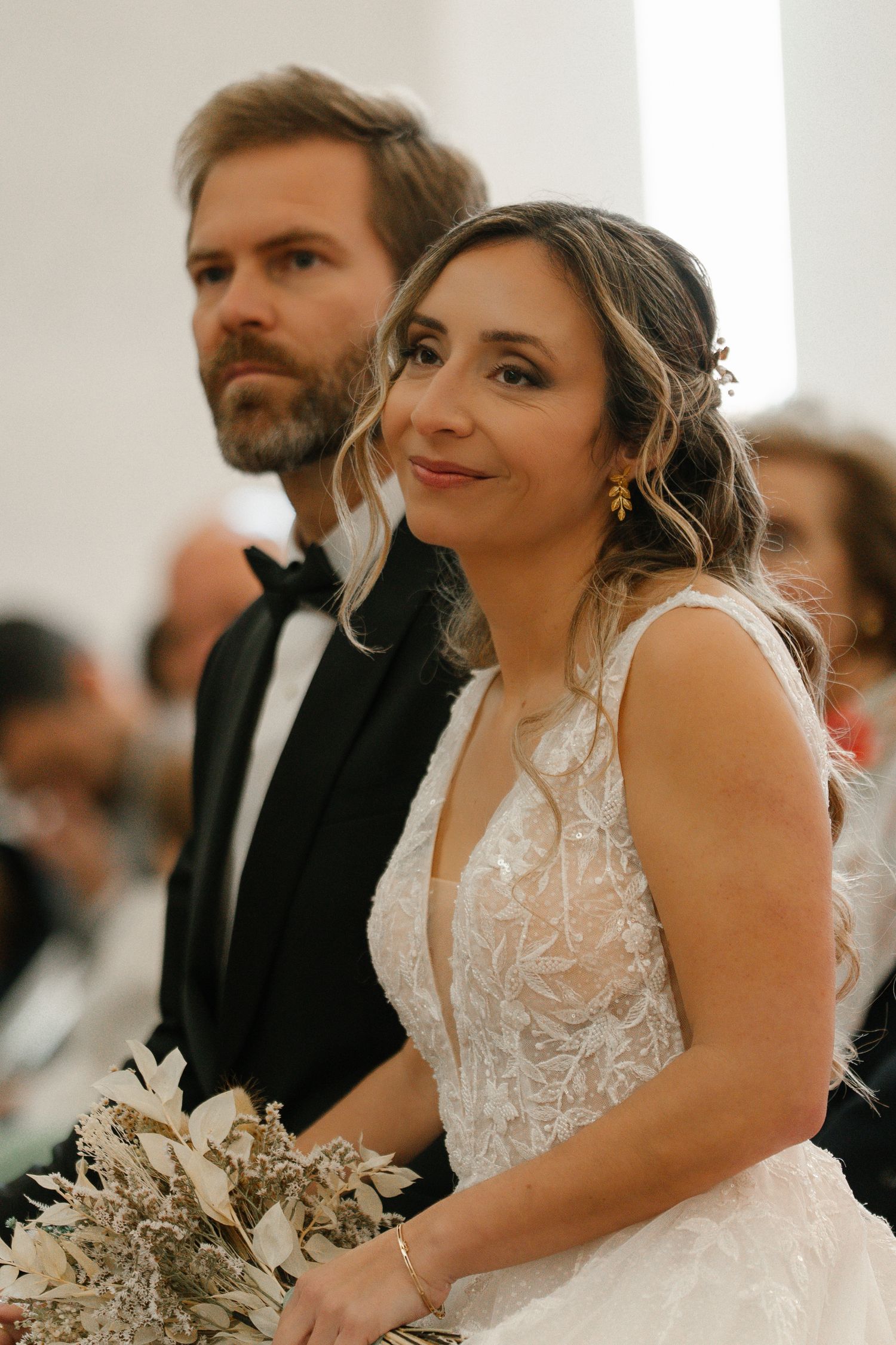 Smiling bride in lace gown holds bouquet beside bearded groom in black tuxedo.