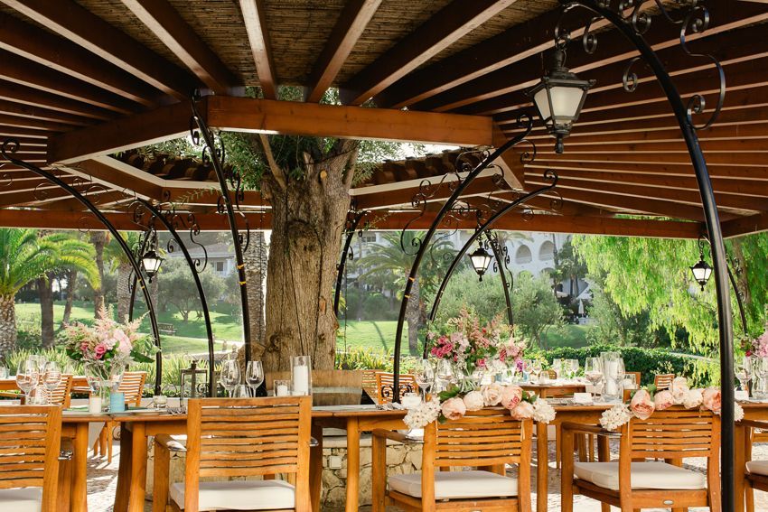 Rustic outdoor wedding reception under wooden pergola with tree centerpiece, wooden tables, and pink floral arrangements.