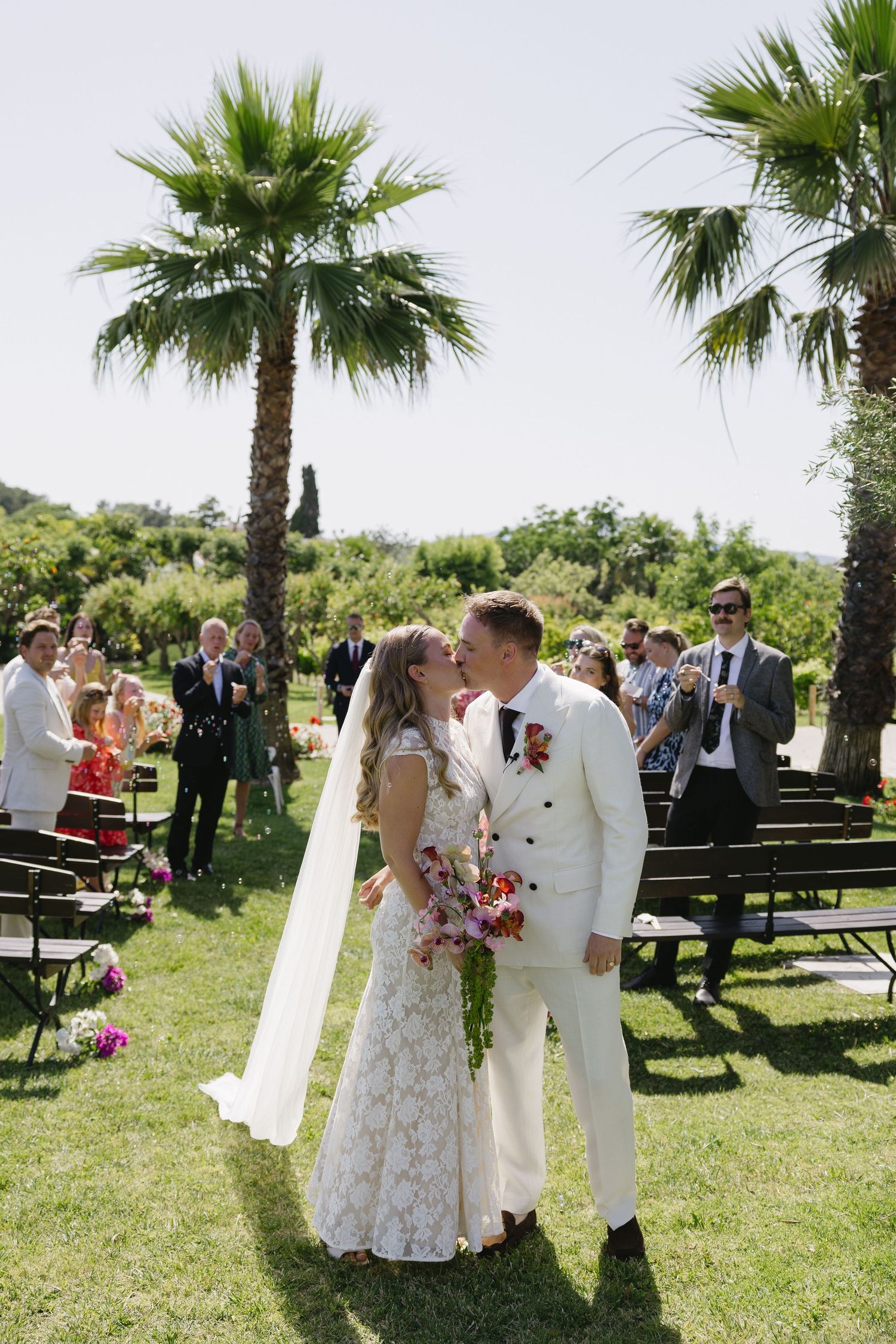 Bride and groom share first kiss at outdoor wedding ceremony with palm trees and guests applauding in sunny garden setting, Algarve Portugal