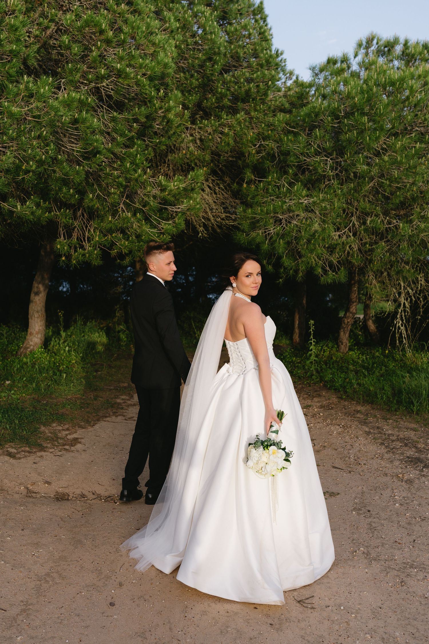 Bride in white ball gown and groom in black tux posing among tall green pine trees.