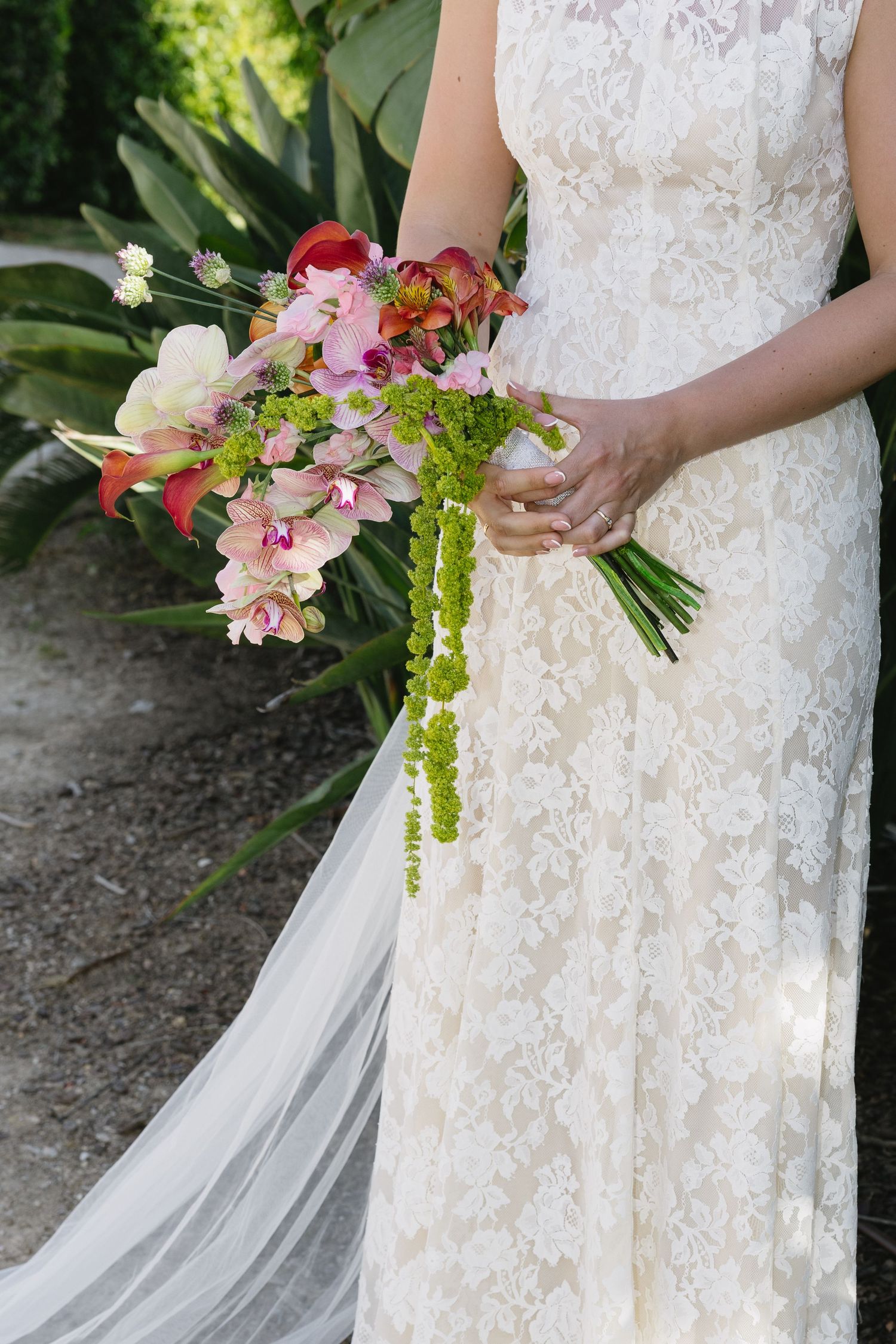 Bride in lace wedding gown holds vibrant tropical bouquet with pink orchids and green hanging amaranthus.