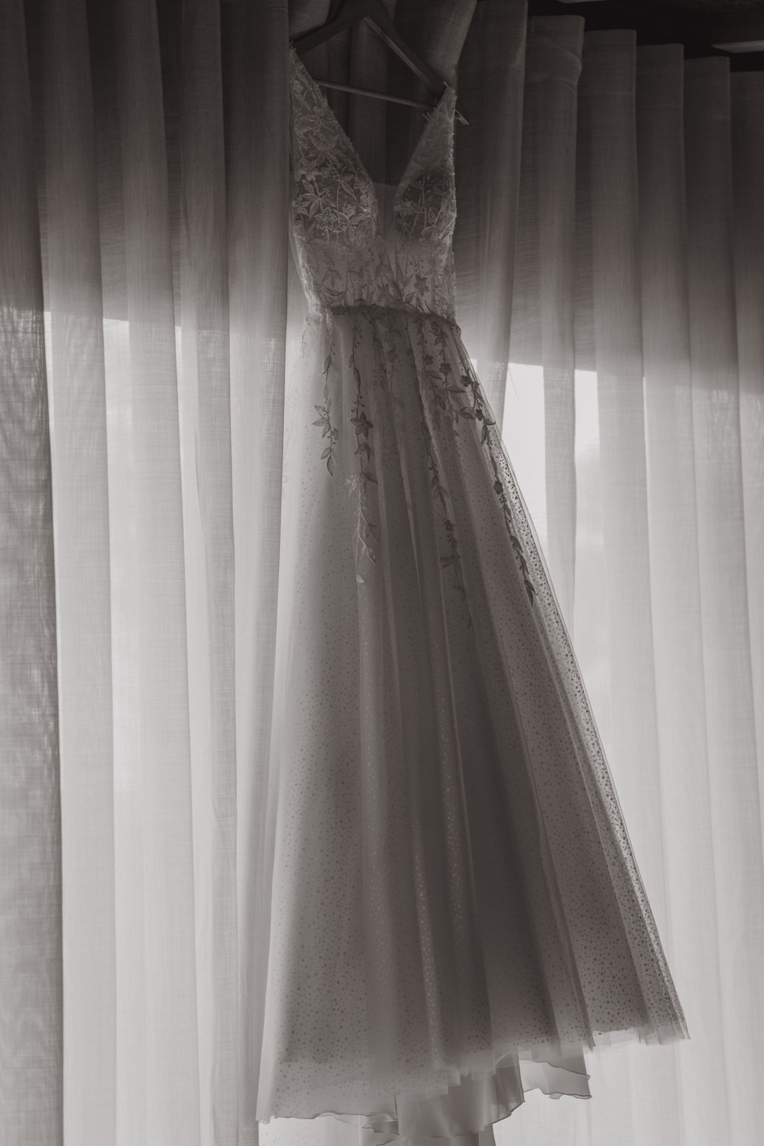 Black and white photo of elegant lace wedding dress hanging by window curtains.