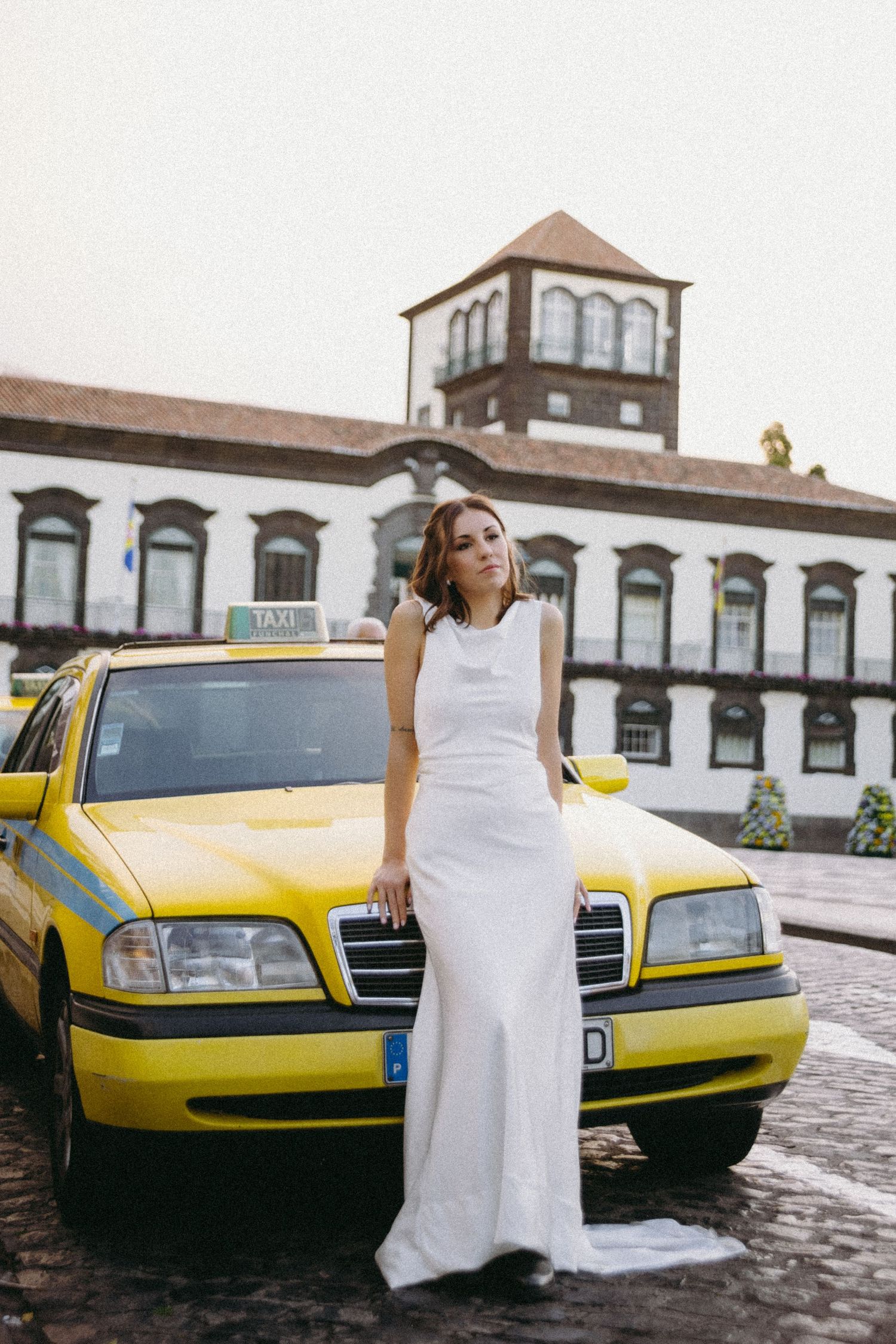 Elegant bride in white gown leans against a yellow taxi cab in front of a historic building.