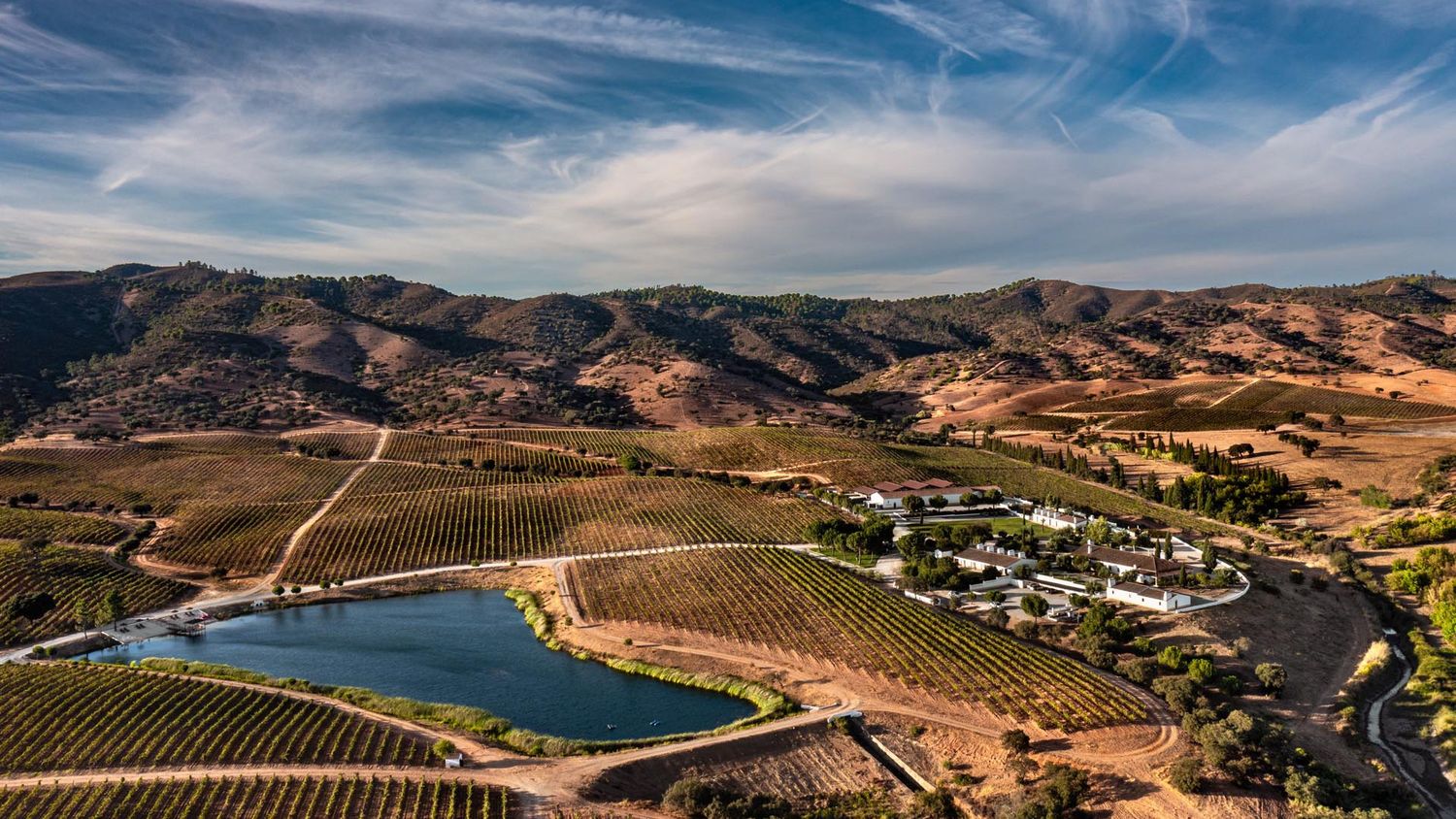 Aerial view of a scenic vineyard estate with a lake, rolling hills, and blue skies.