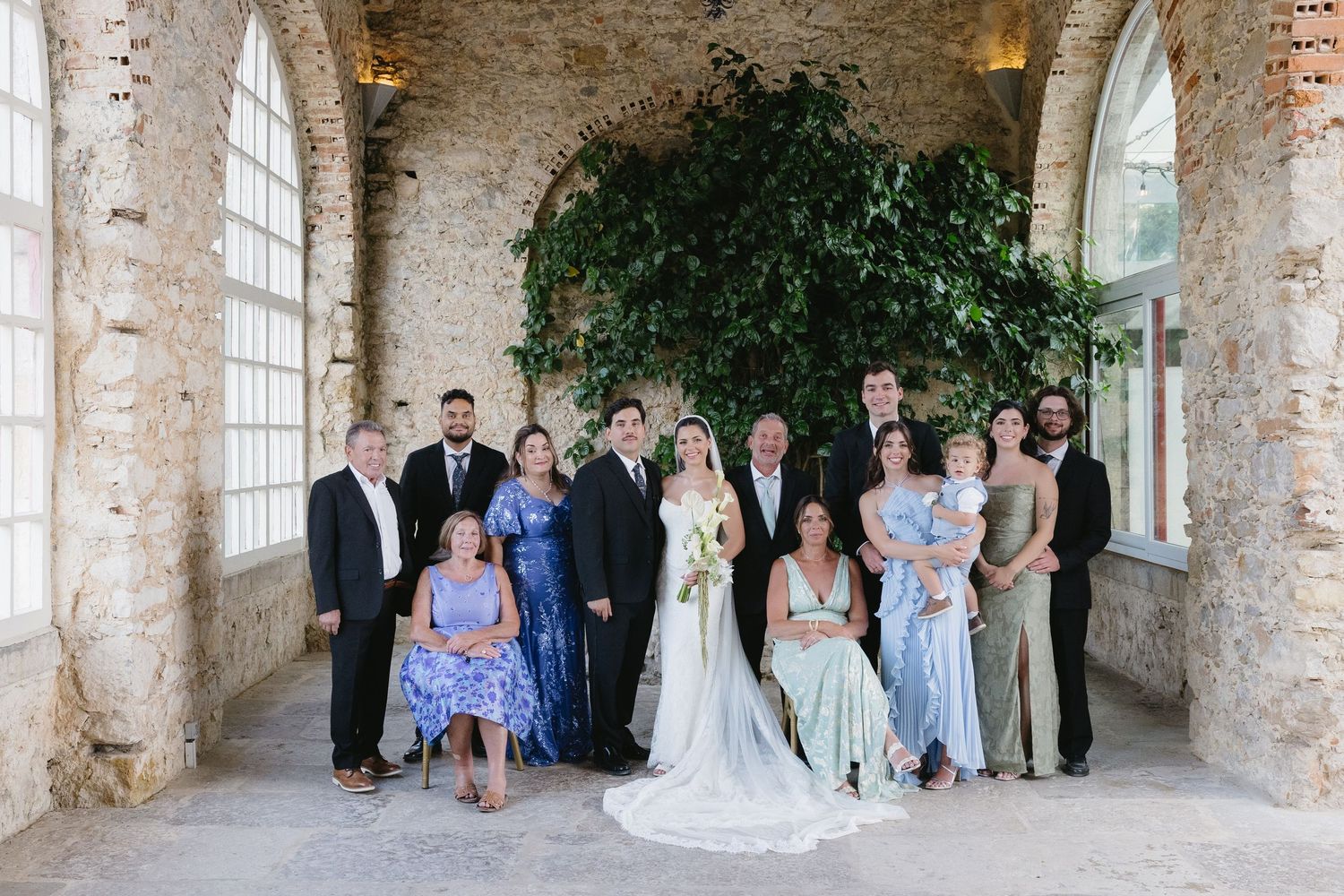 Wedding party poses in elegant stone venue with lush greenery backdrop, bride in white gown surrounded by family in blue dresses.