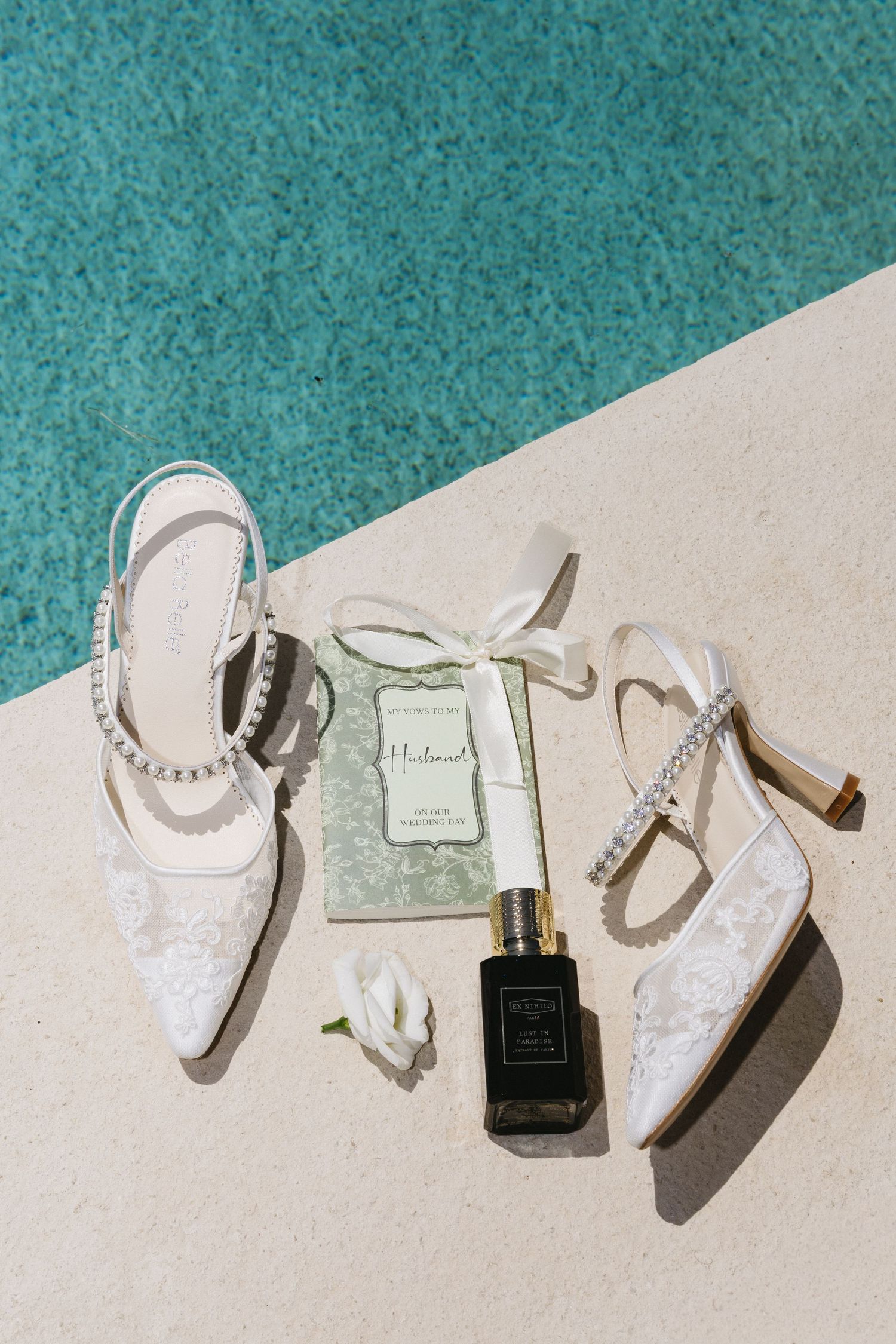 Bridal accessories flatlay featuring white lace heels with crystal straps, perfume bottle, and elegant invitation by poolside.