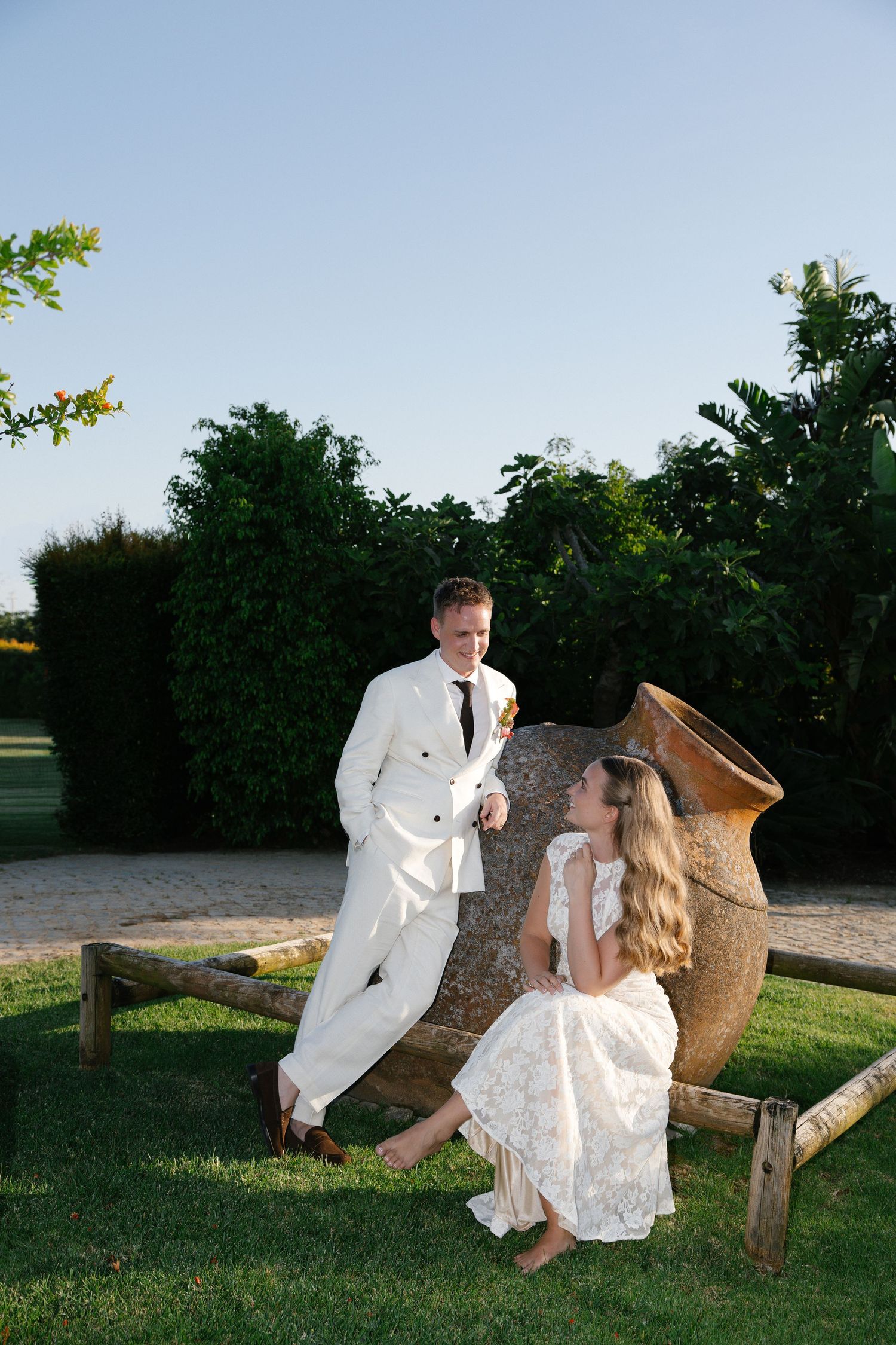 Bride and groom relax beside large terracotta urn in garden at golden hour, white wedding attire.