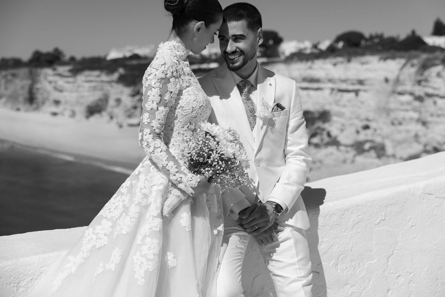 Bride in lace gown and groom in white suit share a tender moment on a coastal cliff in black and white.