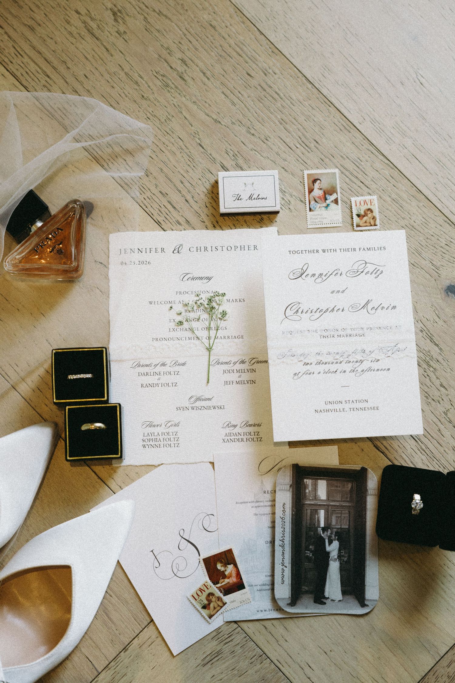 Elegant wedding flat lay with invitations, rings, white heels, perfume, and photos on a wooden floor.