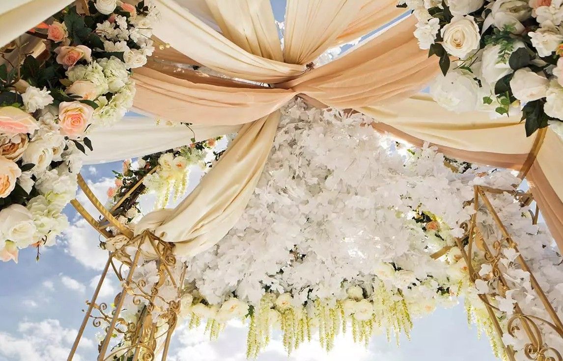 Stunning floral wedding canopy adorned with white flowers and peach draped fabric against blue sky.