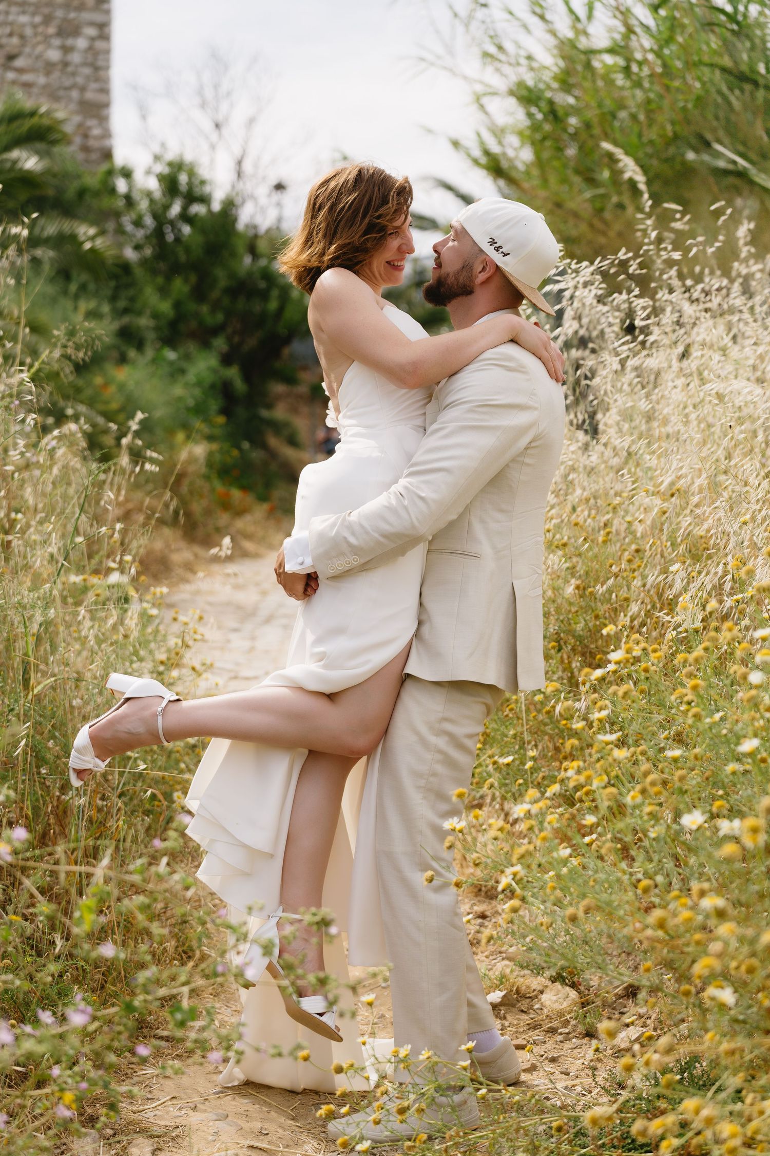 Groom lifts bride among wildflowers in sun-drenched field, sharing a romantic moment.
