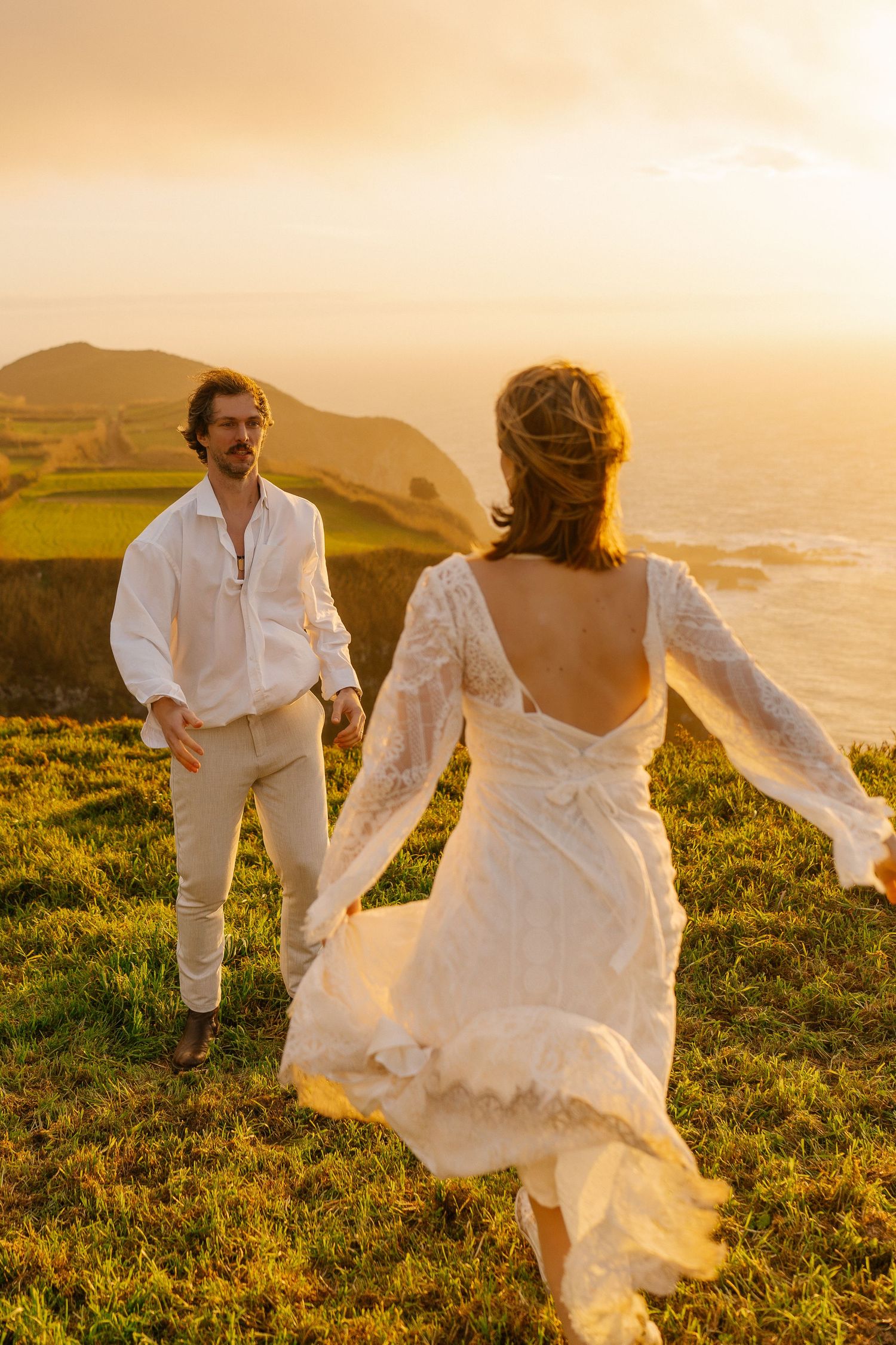 Bride in flowing lace backless gown runs toward groom on golden coastal hillside during stunning sunset elopement.