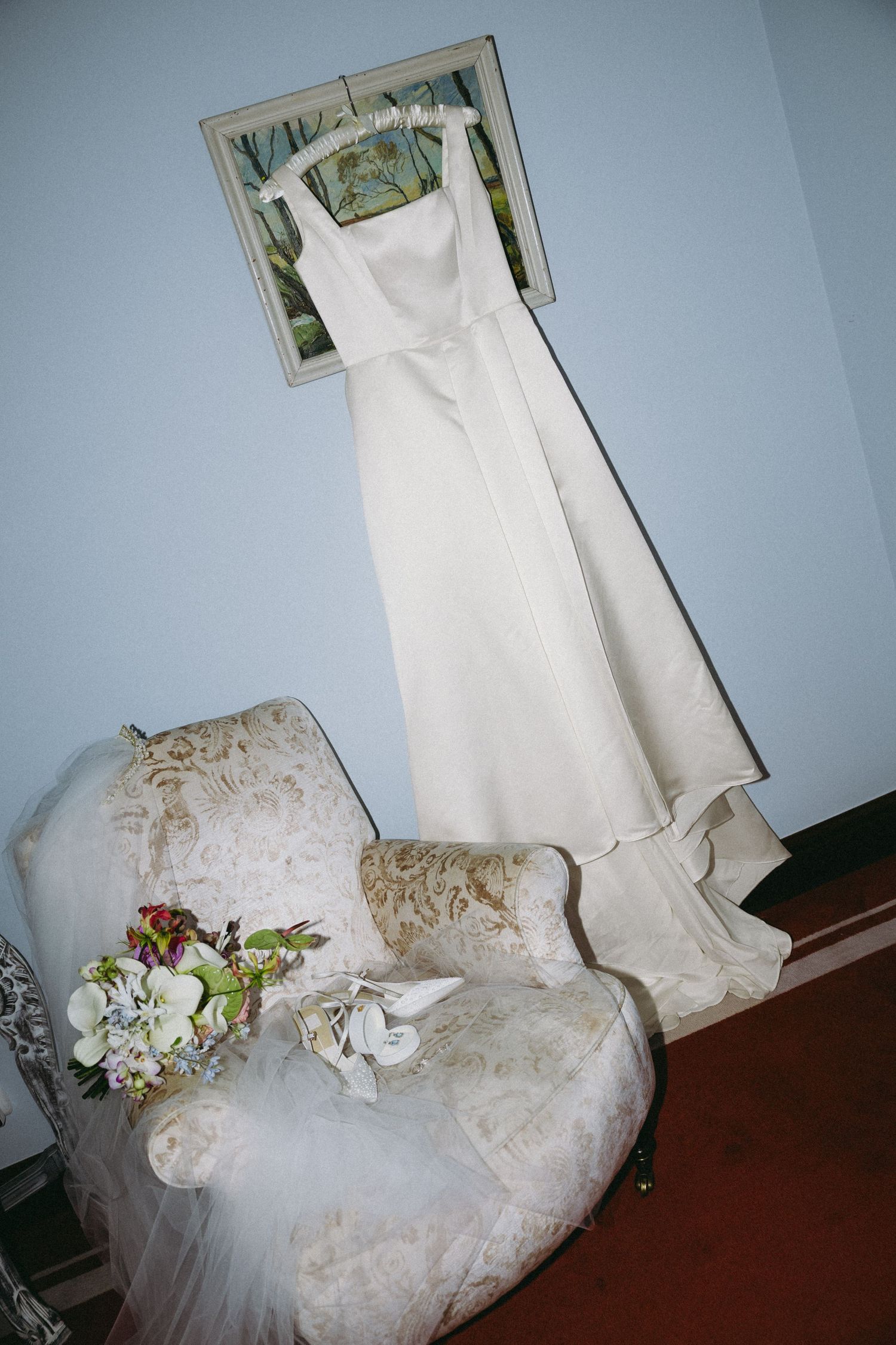 White wedding dress hanging on wall above floral chair with bridal bouquet and heels displayed below.