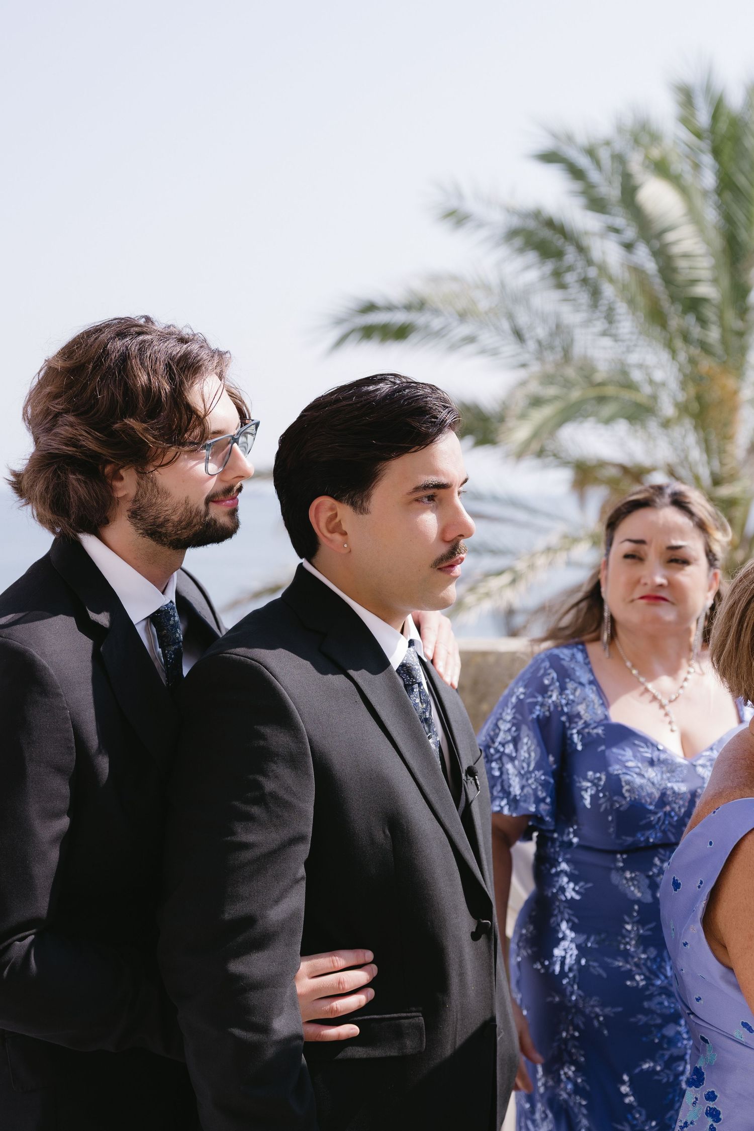 Two men in black suits and a woman in blue dress stand outdoors near palm trees at a formal event.