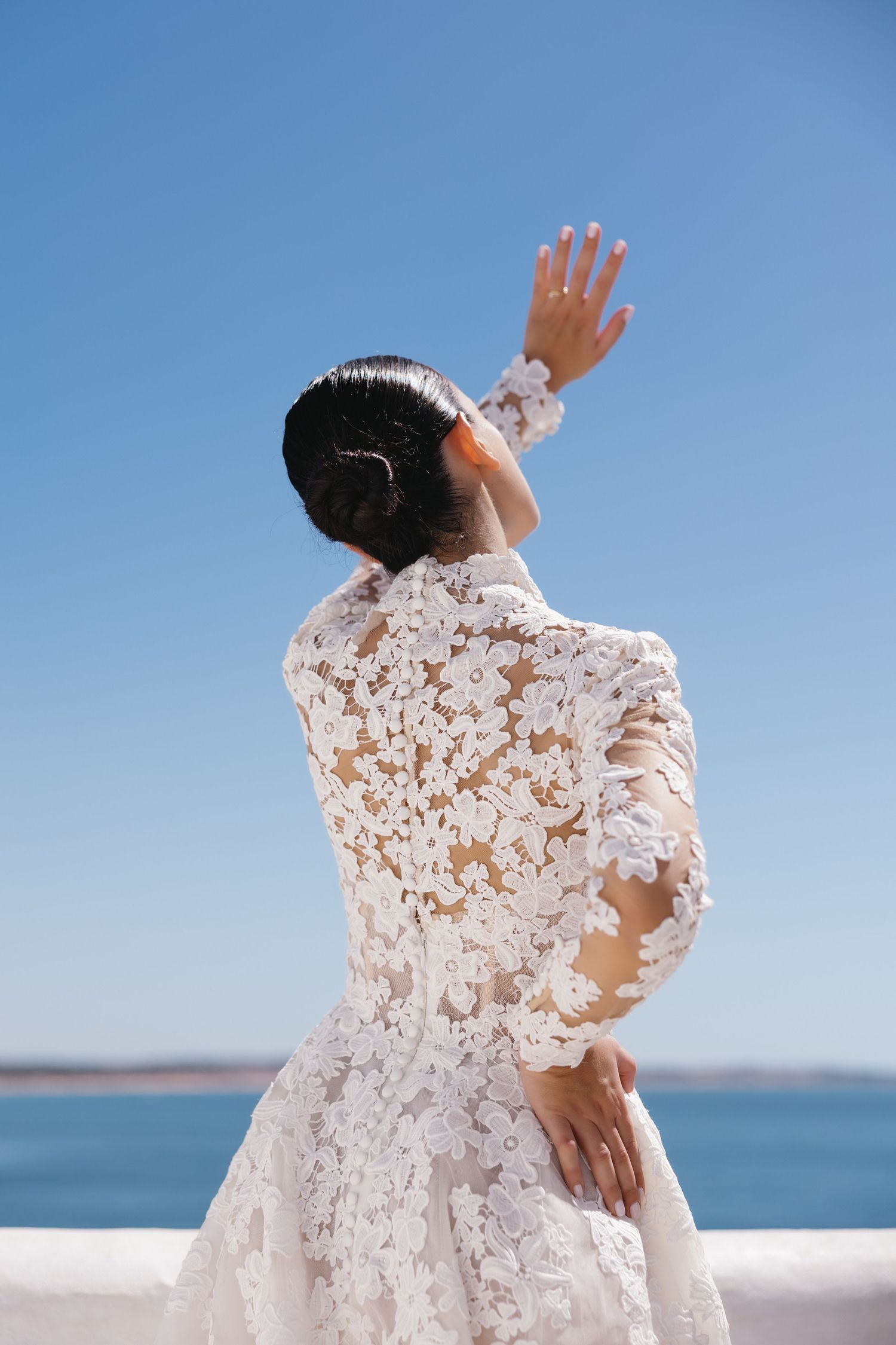 Bride in elegant floral lace wedding dress raises hand against clear blue sky by the sea.