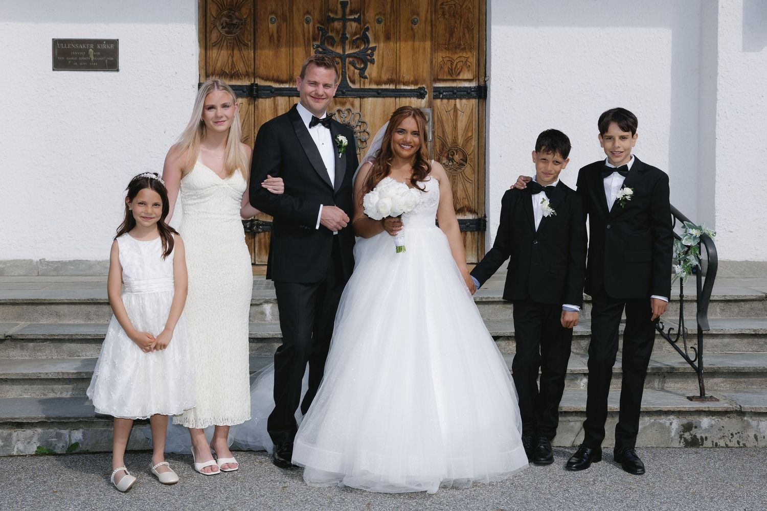A bride and groom pose with three children in formal attire outside a church on their wedding day.