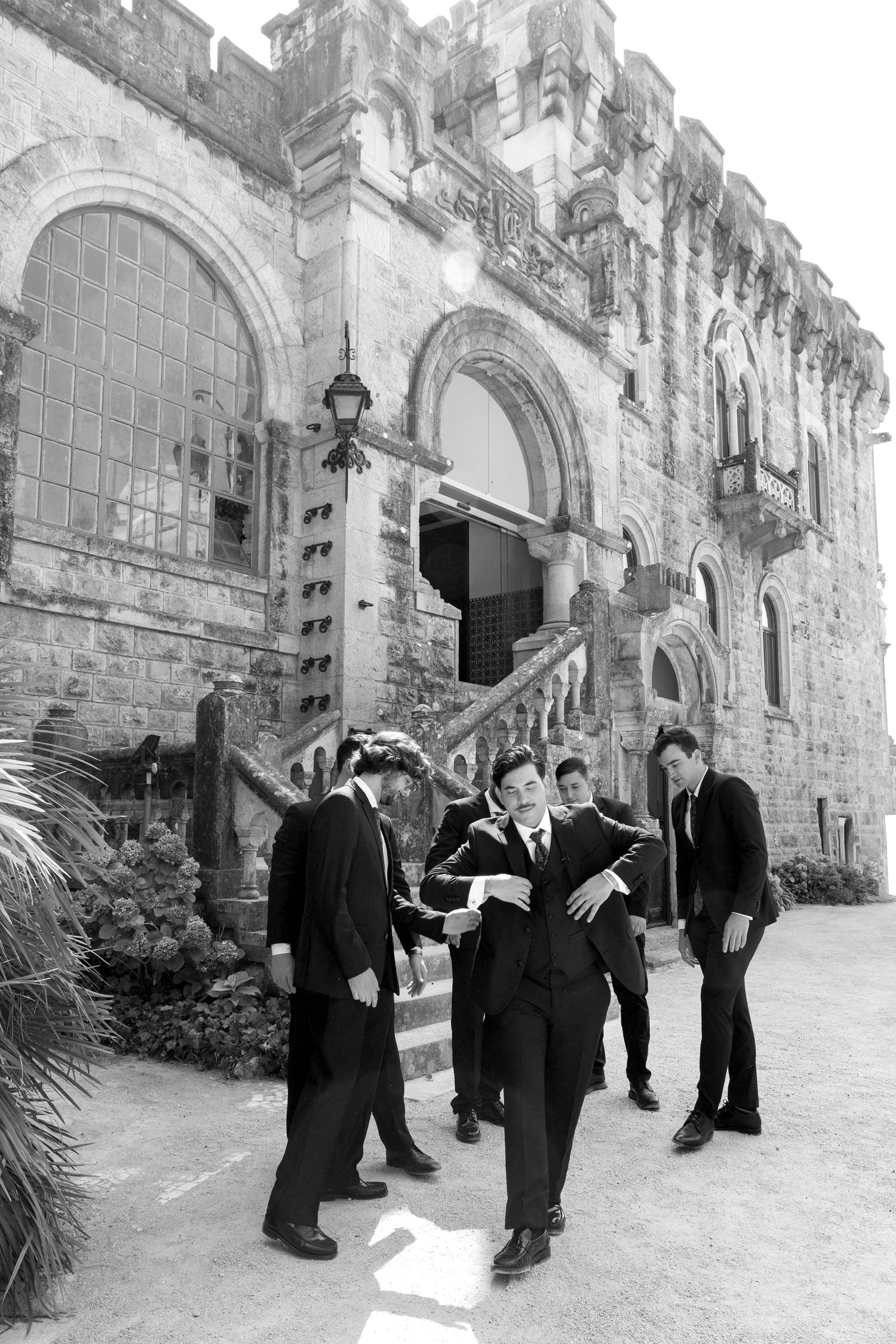 Black and white photo of groomsmen in suits gathered outside a historic stone building with ornate architecture.