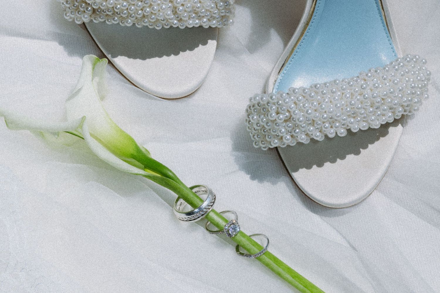 Pearl-embellished white bridal heels beside a calla lily and diamond ring on white fabric.