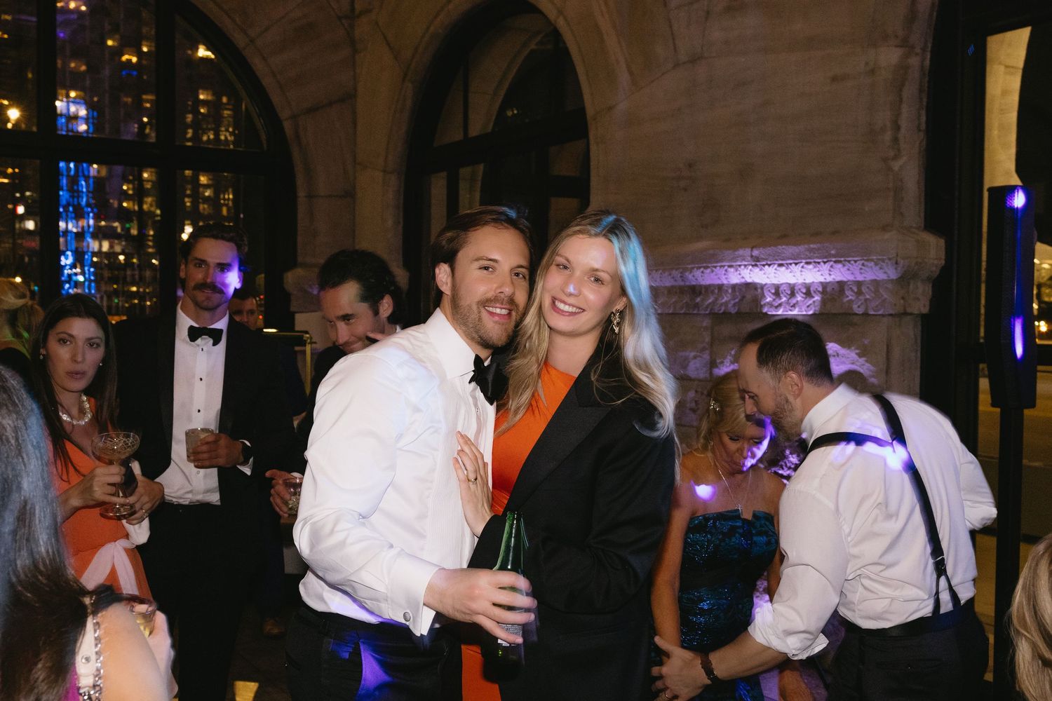 A man and woman smile together at an elegant evening gala event with purple lighting and guests in formal attire.