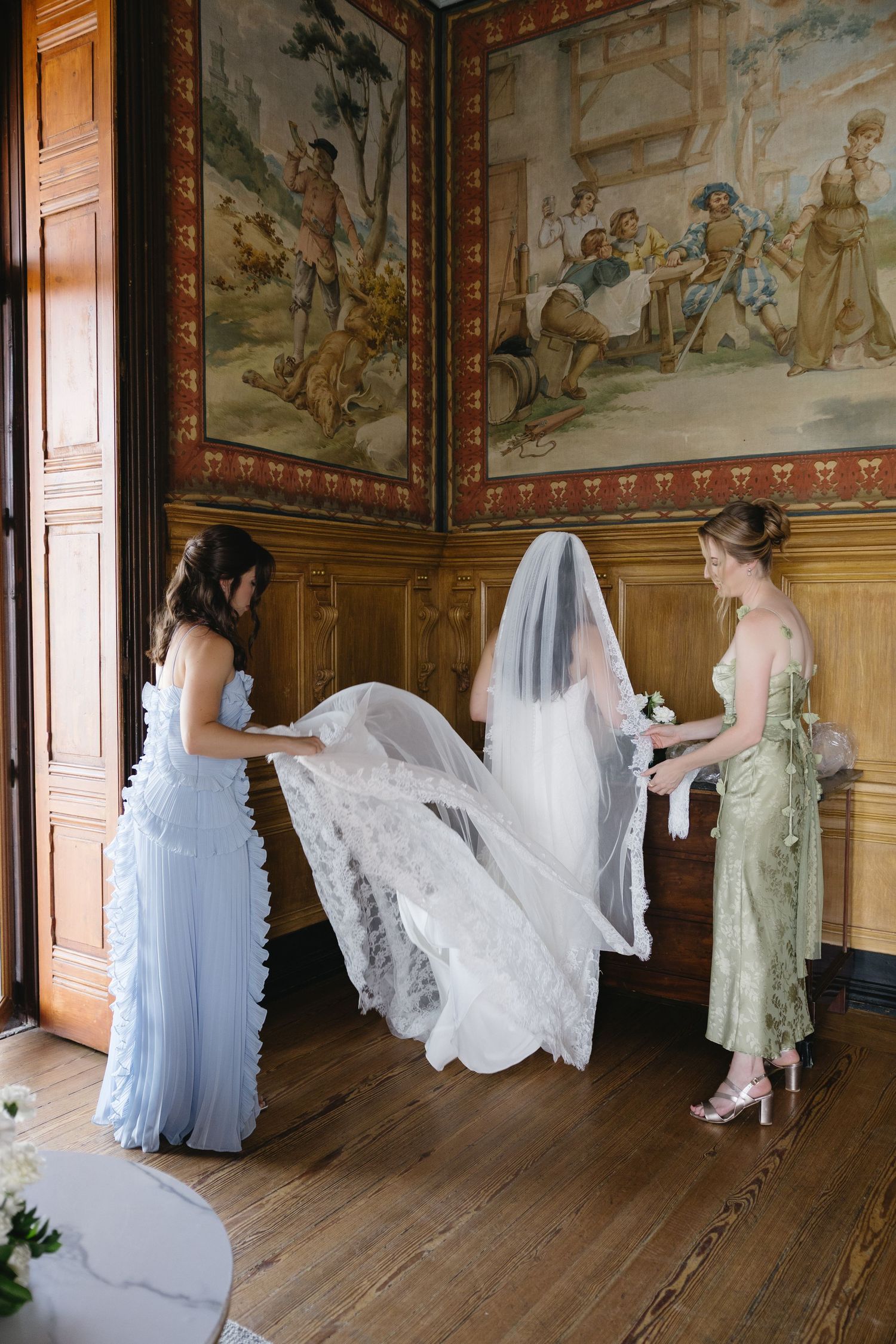 Two bridesmaids in blue and green gowns arrange a bride's lace veil in an ornate wood-paneled room with tapestries.
