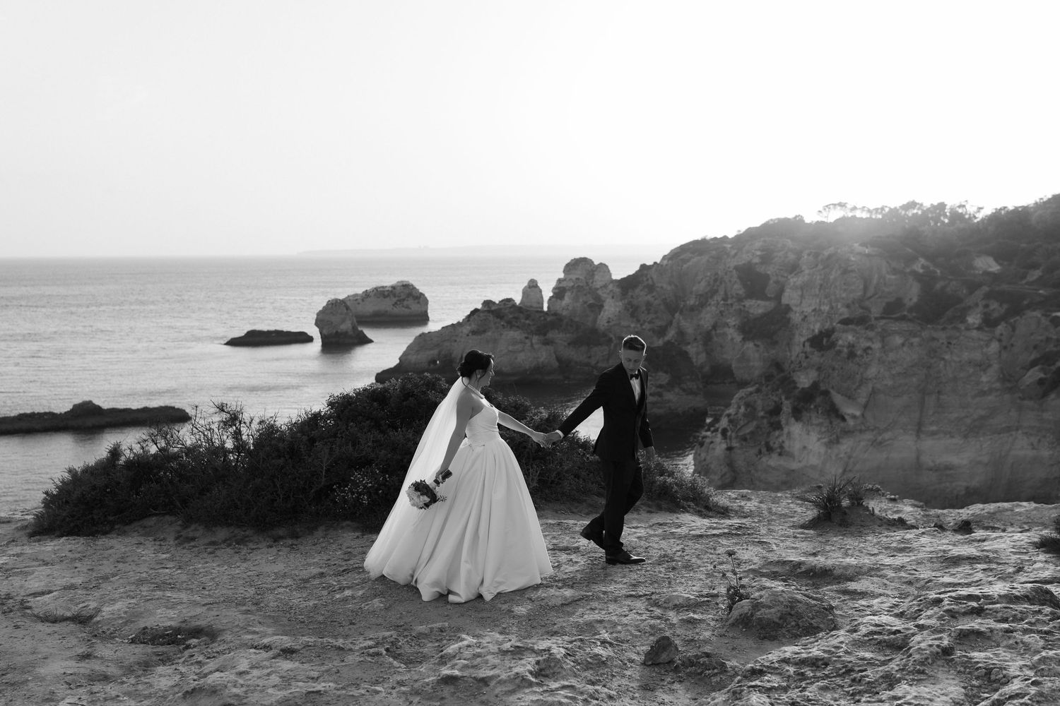 Black and white photo of bride and groom holding hands walking along dramatic ocean cliffs.