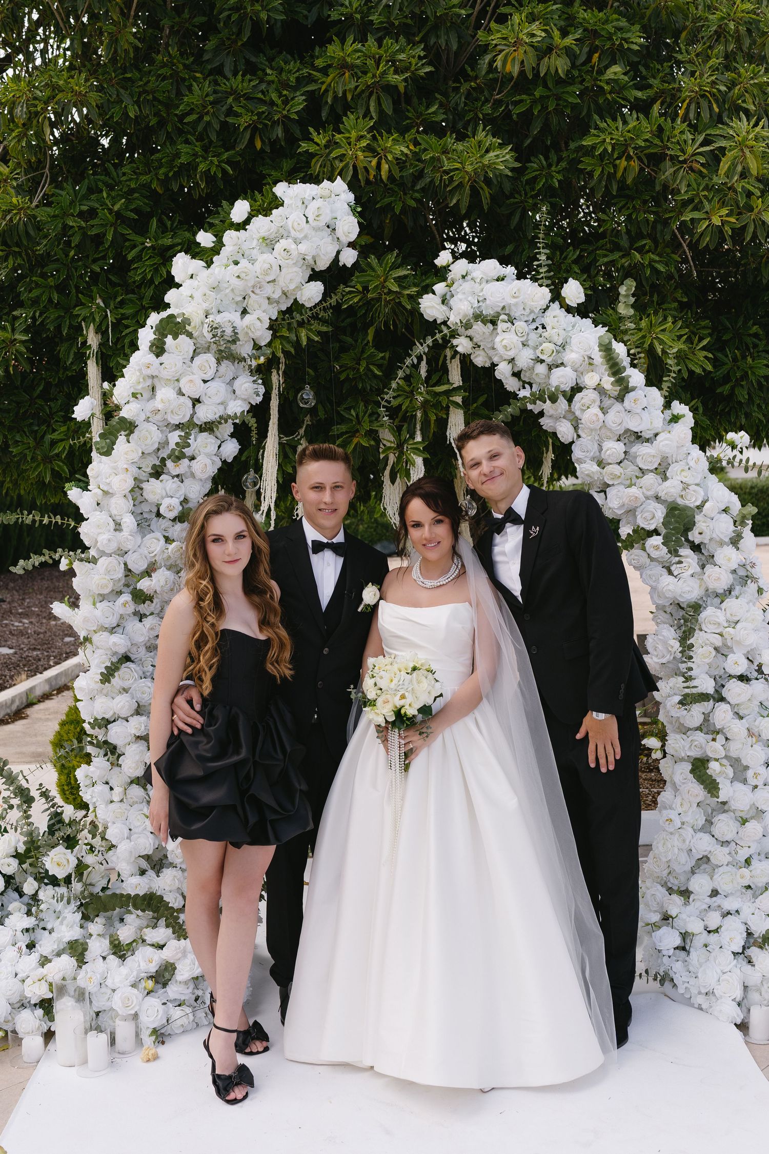 Wedding party posing under white floral arch with bride, groom, and two guests in formal attire.