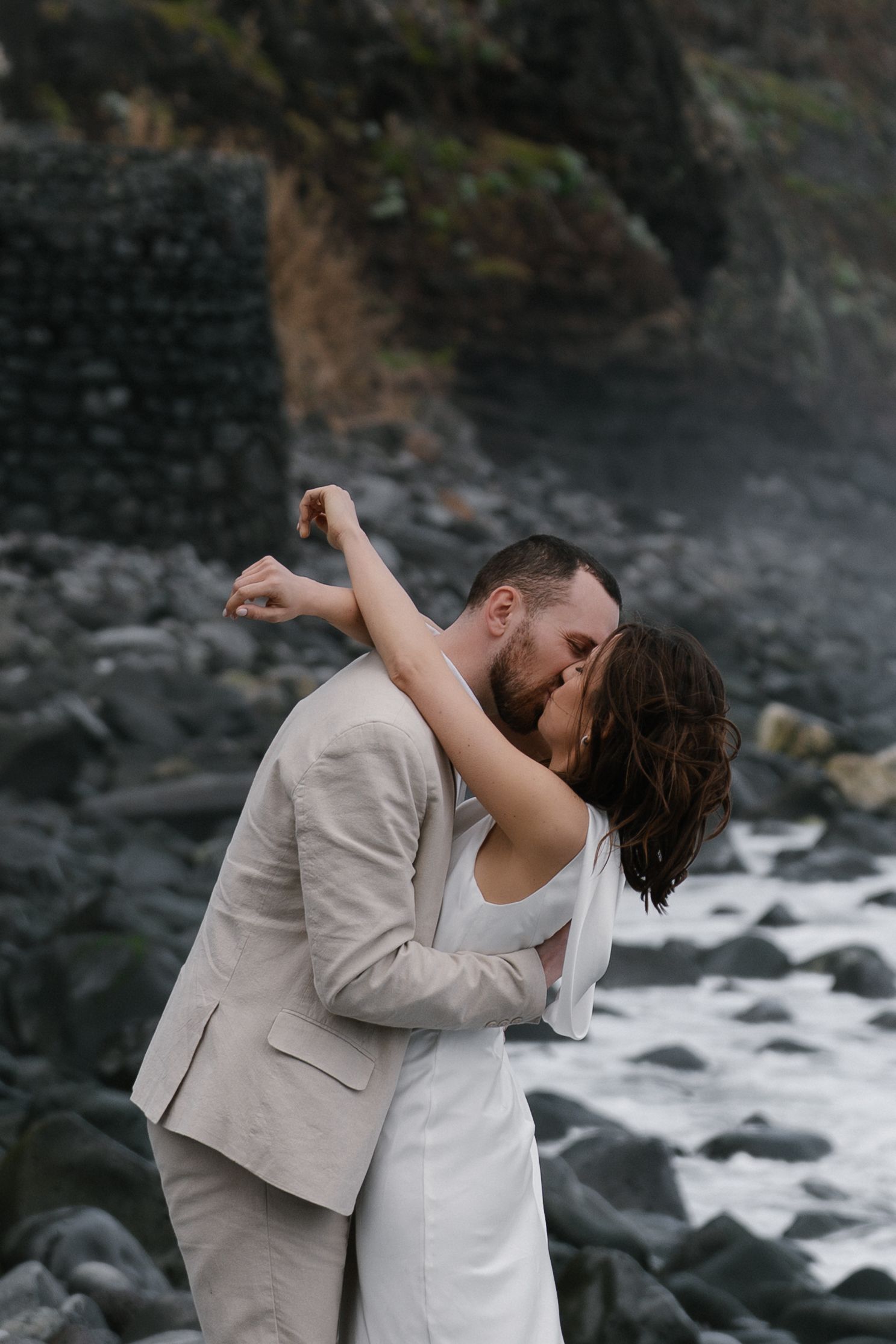 Couple sharing a passionate kiss on a rocky beach with crashing waves.