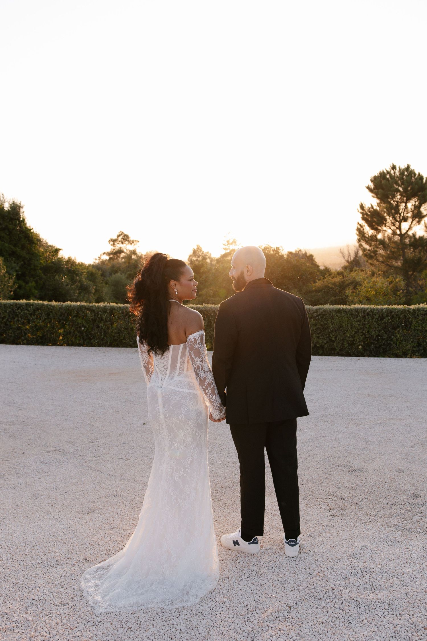 Bride and groom holding hands at sunset, viewed from behind, bride in lace off-shoulder gown on gravel path.