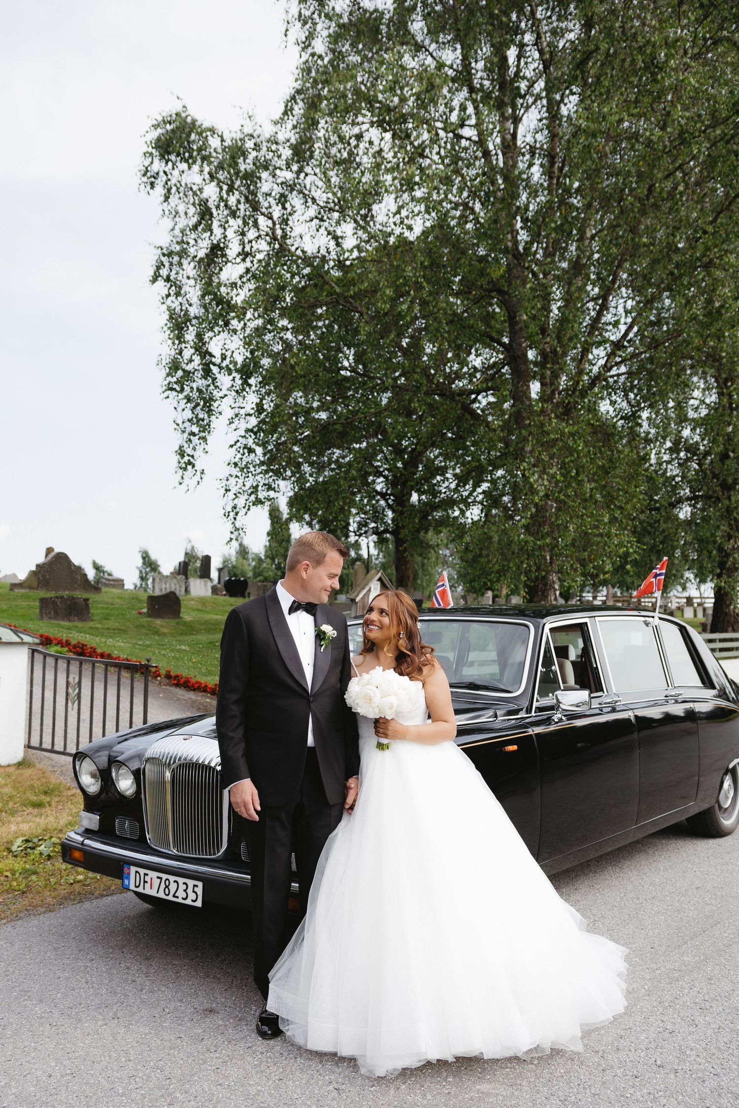 Bride and groom pose beside a black vintage limousine with Norwegian flags, surrounded by lush green trees.