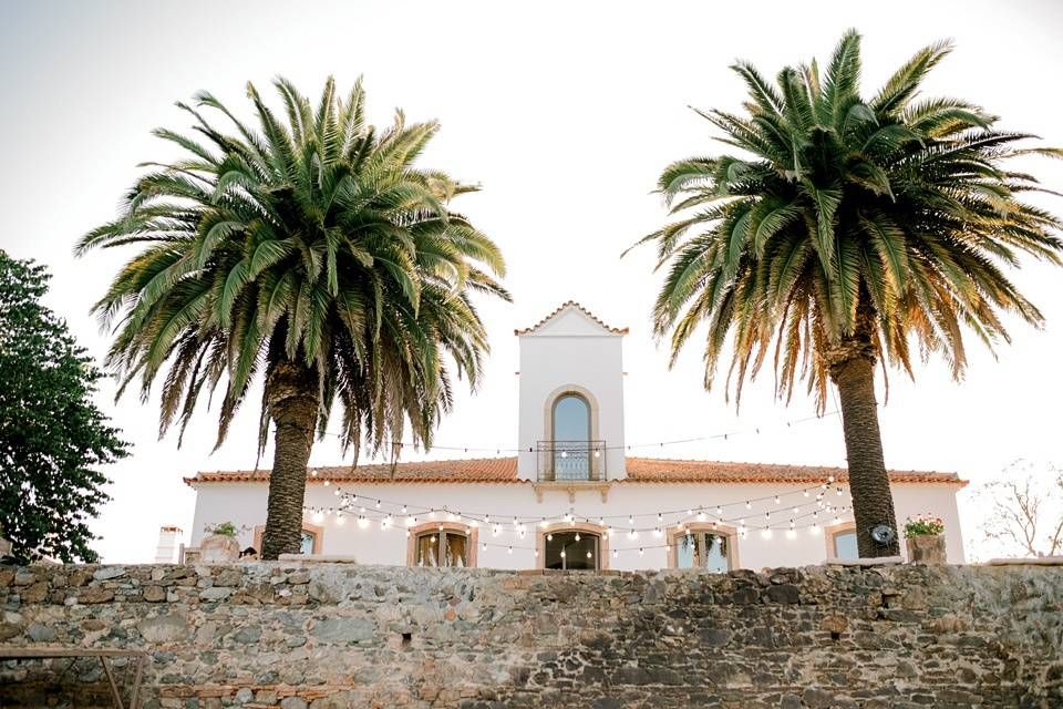White Spanish-style wedding venue with red tile roof framed by two tall palm trees under an overcast sky.