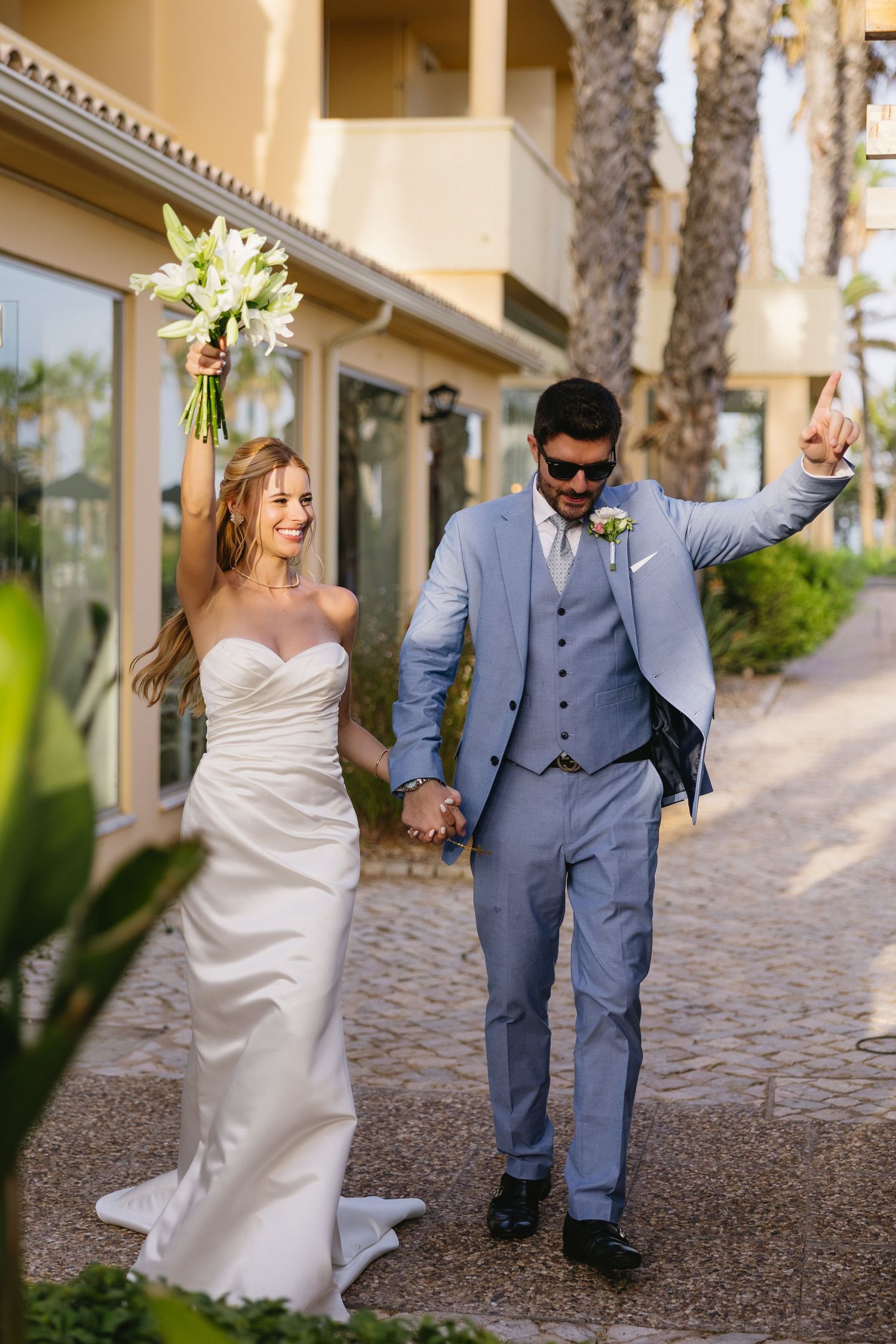 Joyful newlyweds walk hand-in-hand, bride raising bouquet, groom in light blue suit, celebrating after outdoor wedding ceremony.