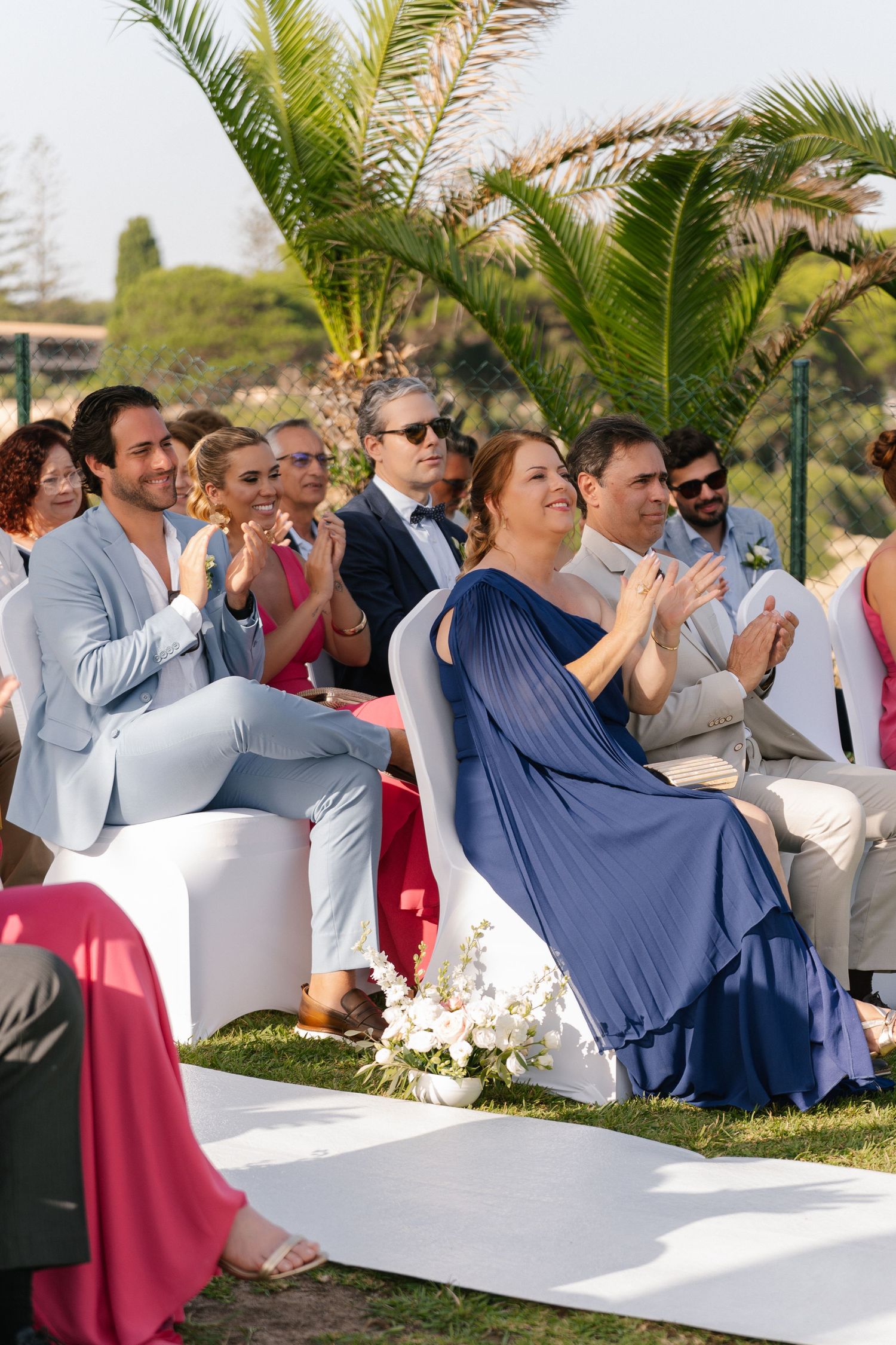 Wedding guests in elegant attire applaud joyfully during an outdoor ceremony surrounded by lush greenery.